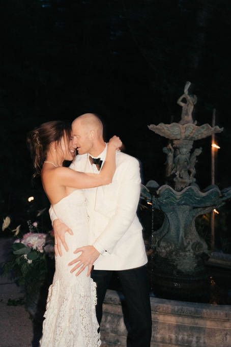A romantic wedding couple shares a kiss at night beside an ornate fountain with decorative cross details.