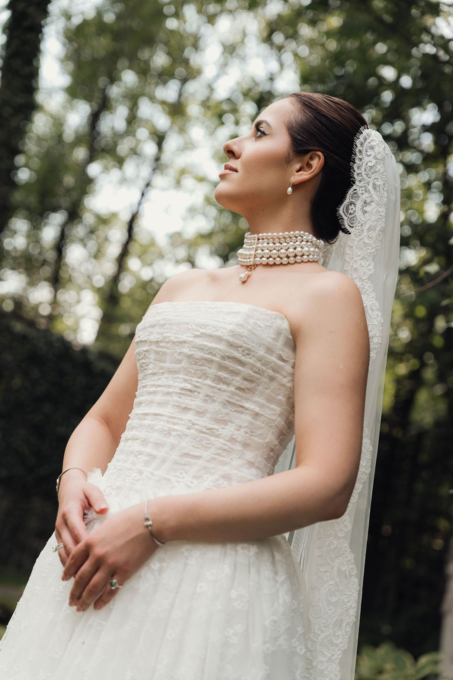 Elegant bride in strapless white lace gown with pearl necklace and veil poses outdoors among lush green trees.