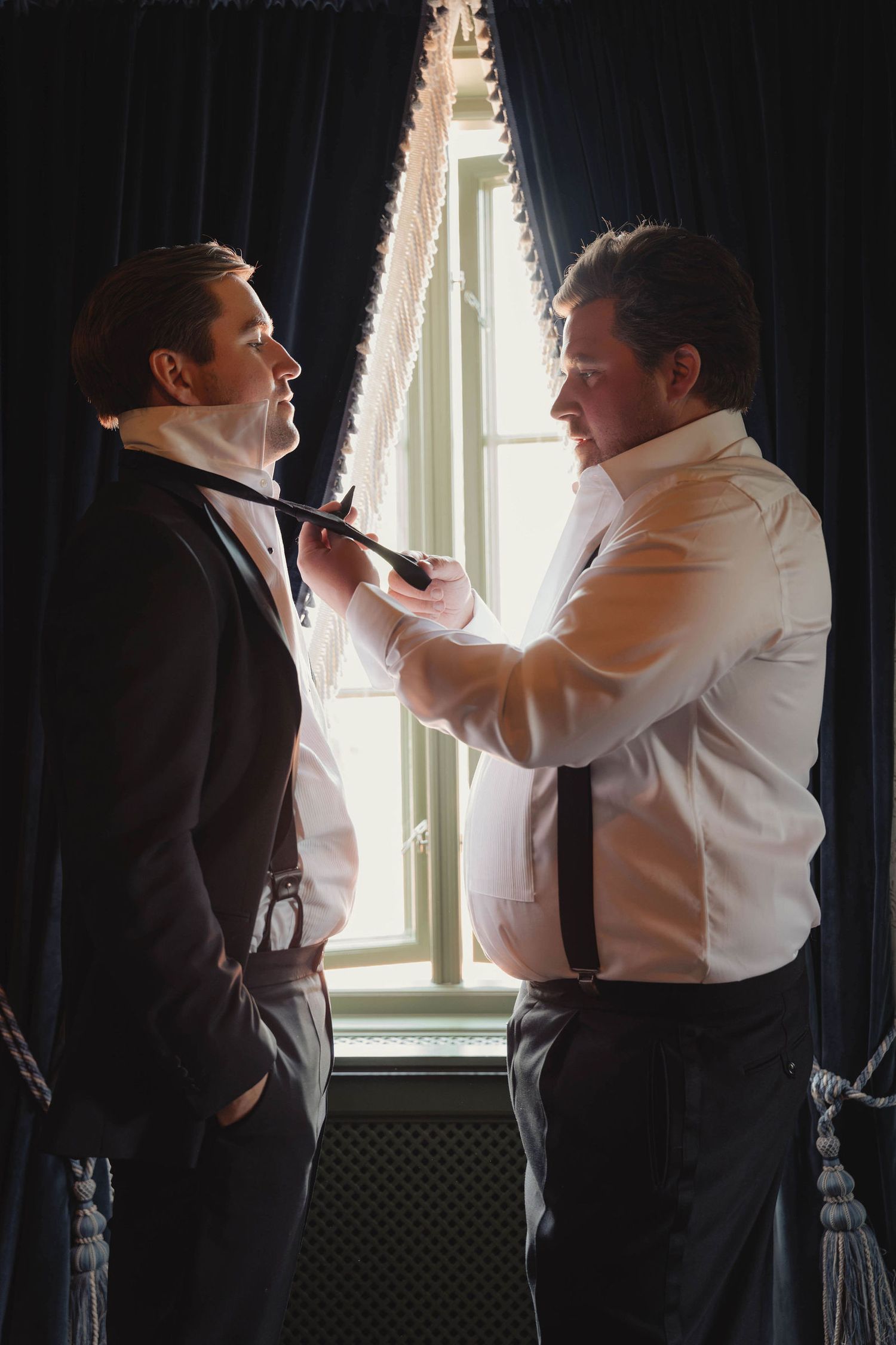 Two men in formal attire help each other with neckties near a window with dark curtains.