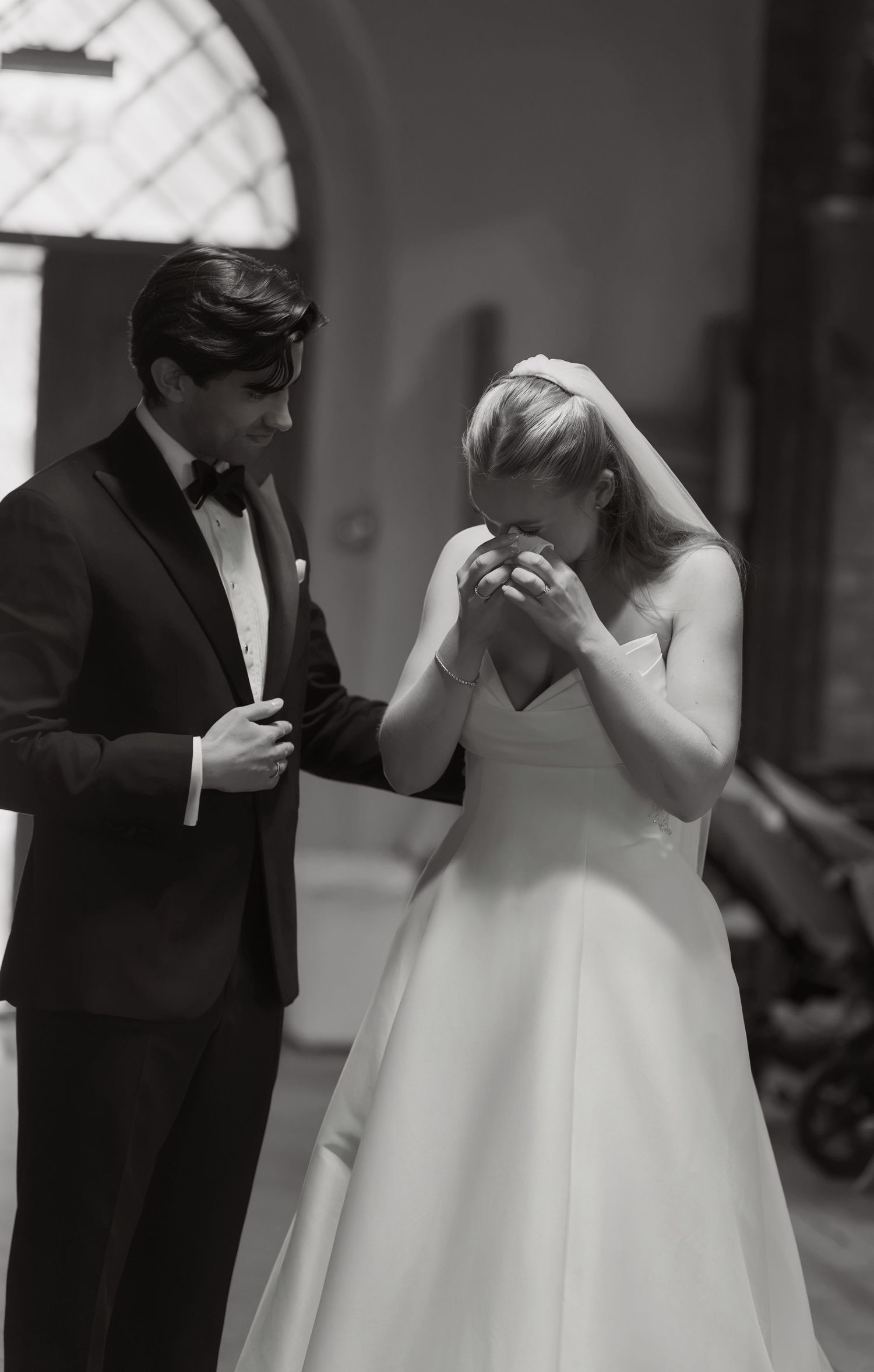 Black and white photo of a bride in a strapless gown and veil covering her face with emotion as the groom in a tuxedo looks on.