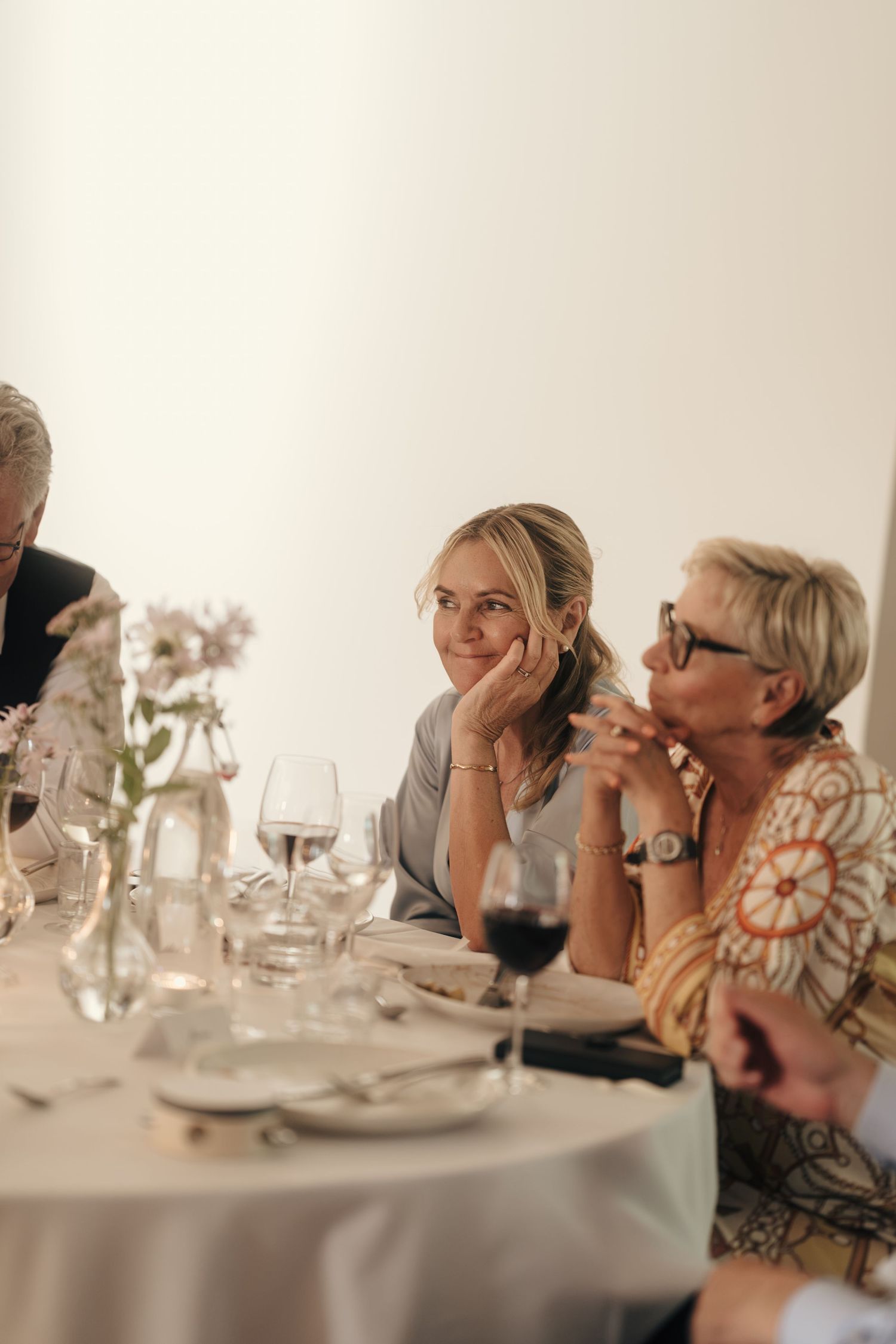 Two smiling women lean on a white-clothed table set with wine glasses and flowers during a wedding reception dinner.