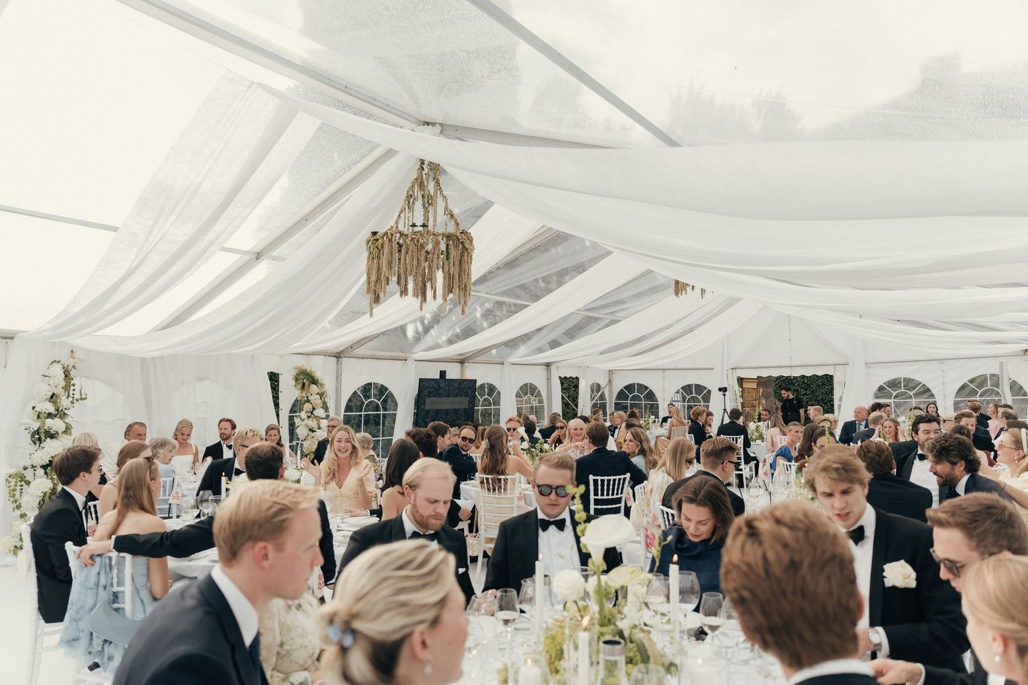Wedding guests in formal attire seated at long tables under a white draped marquee tent with a chandelier during a reception dinner.