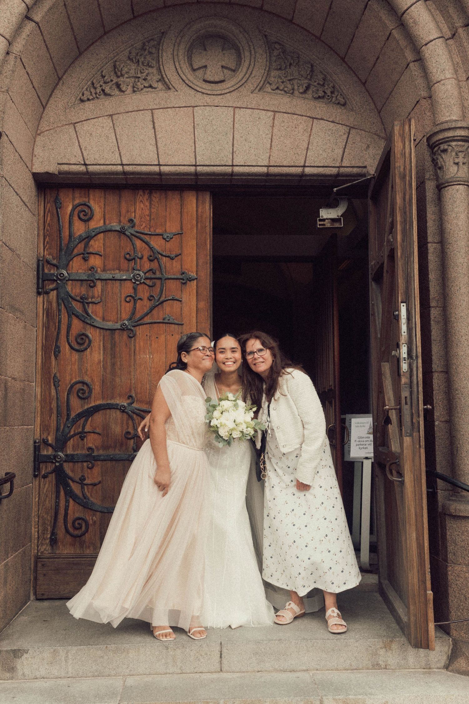 Three people in white dresses sit together on stone church steps with ornate wooden doors in the background.