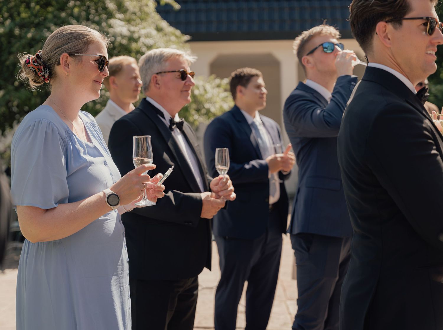Wedding guests in formal attire, including a woman in a light blue dress, hold champagne glasses while standing outdoors in a garden.