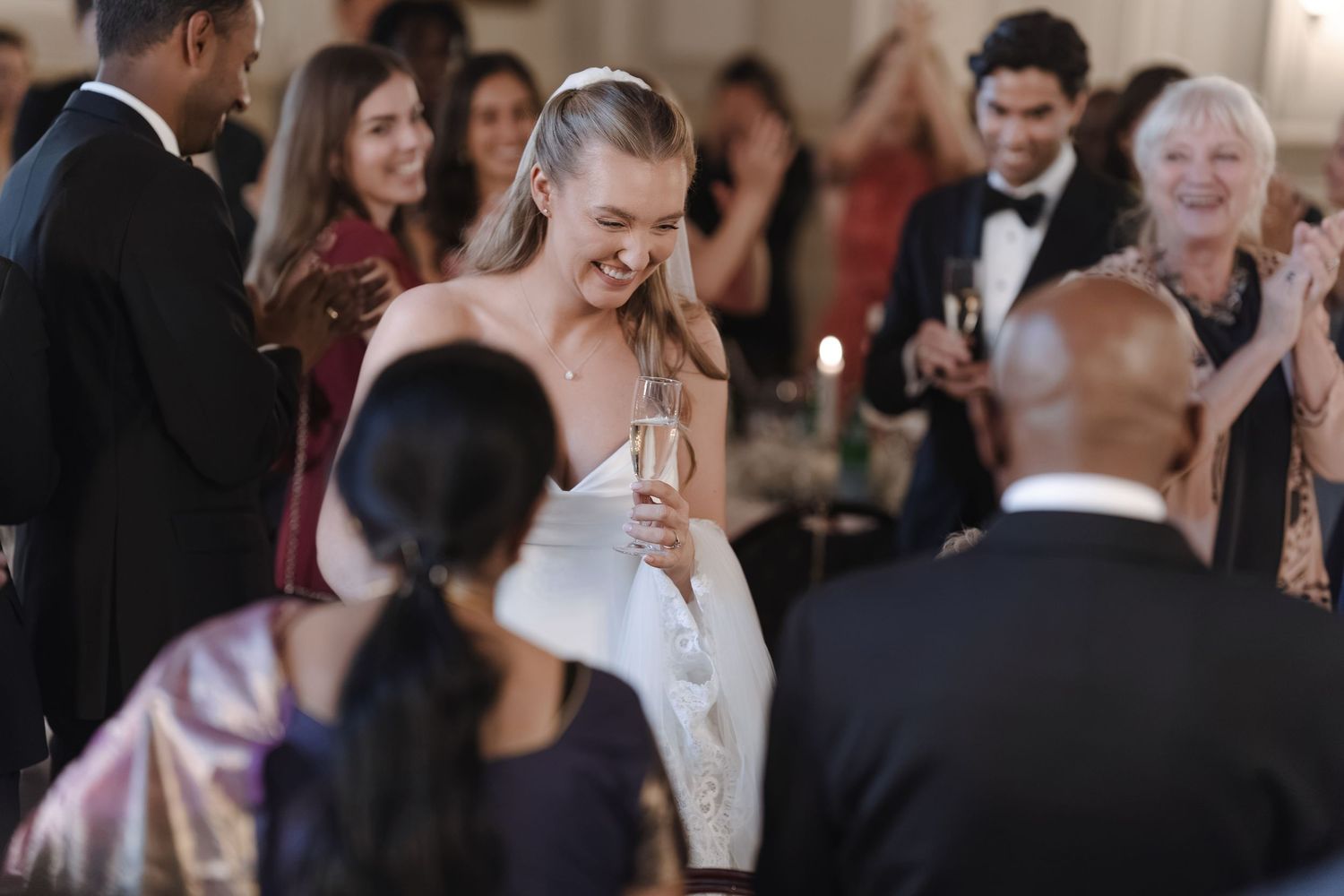Smiling bride in a white strapless gown holds a champagne flute while wedding guests in formal attire applaud and celebrate around her.