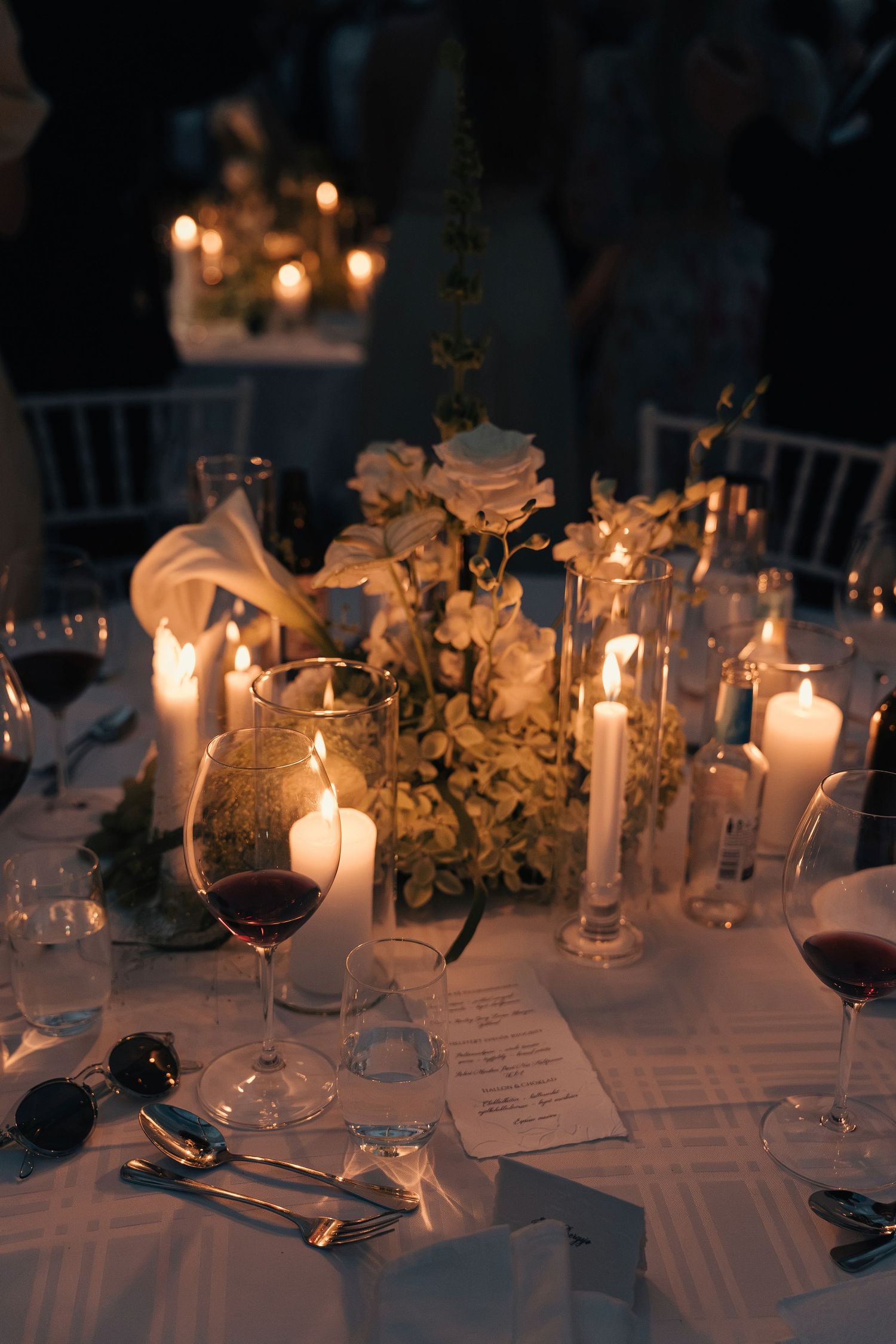 Candlelit wedding reception table with white roses, red wine glasses, and a printed menu card in warm light.