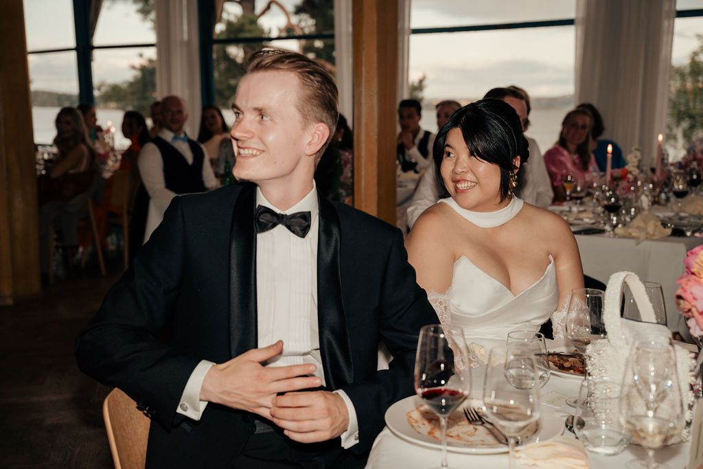 Elegantly dressed couple smiles while seated at a formal reception dinner table with wine glasses and place settings.