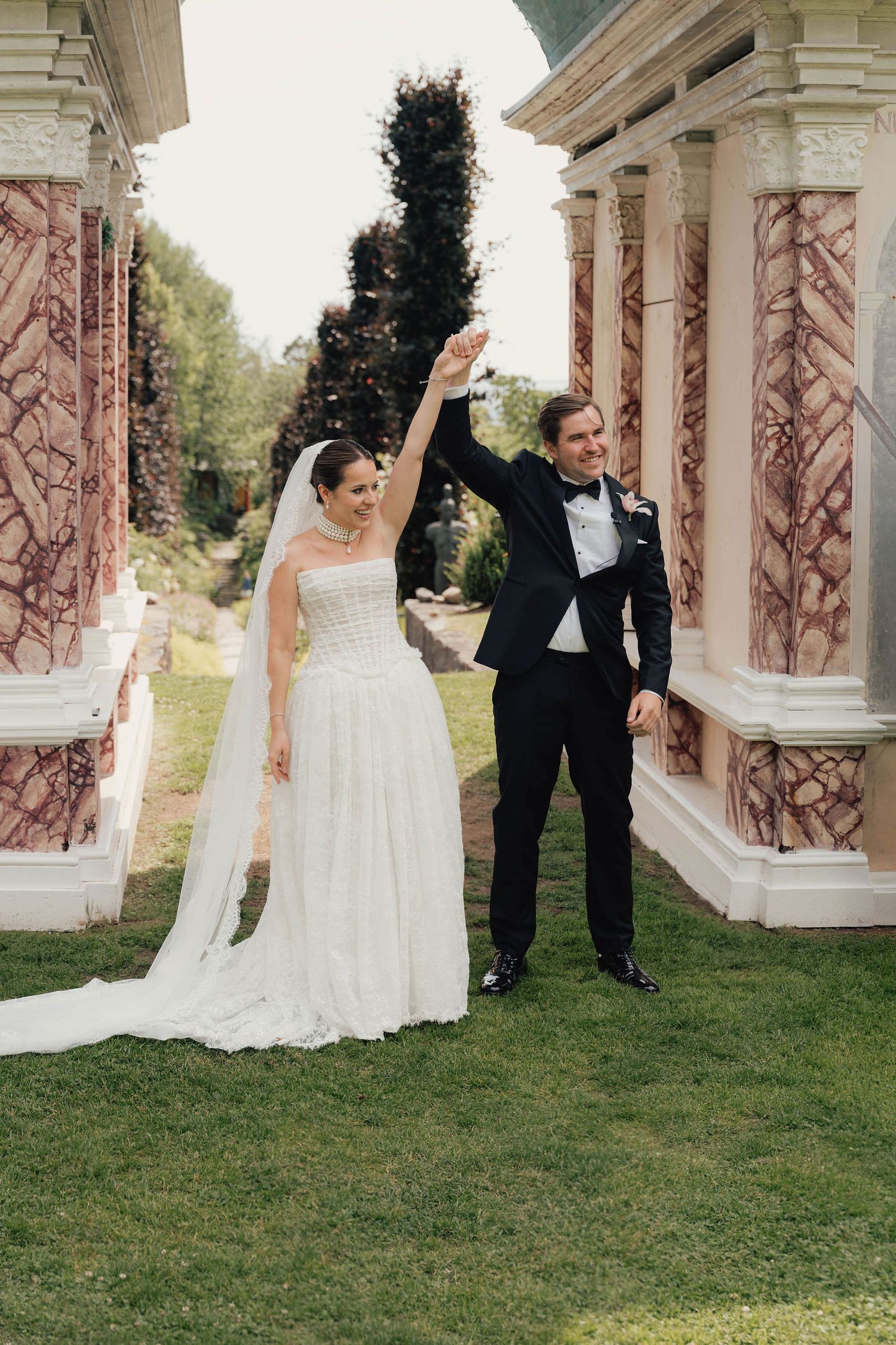 Bride and groom celebrate with raised hands beside elegant marble columns in a lush garden wedding setting.