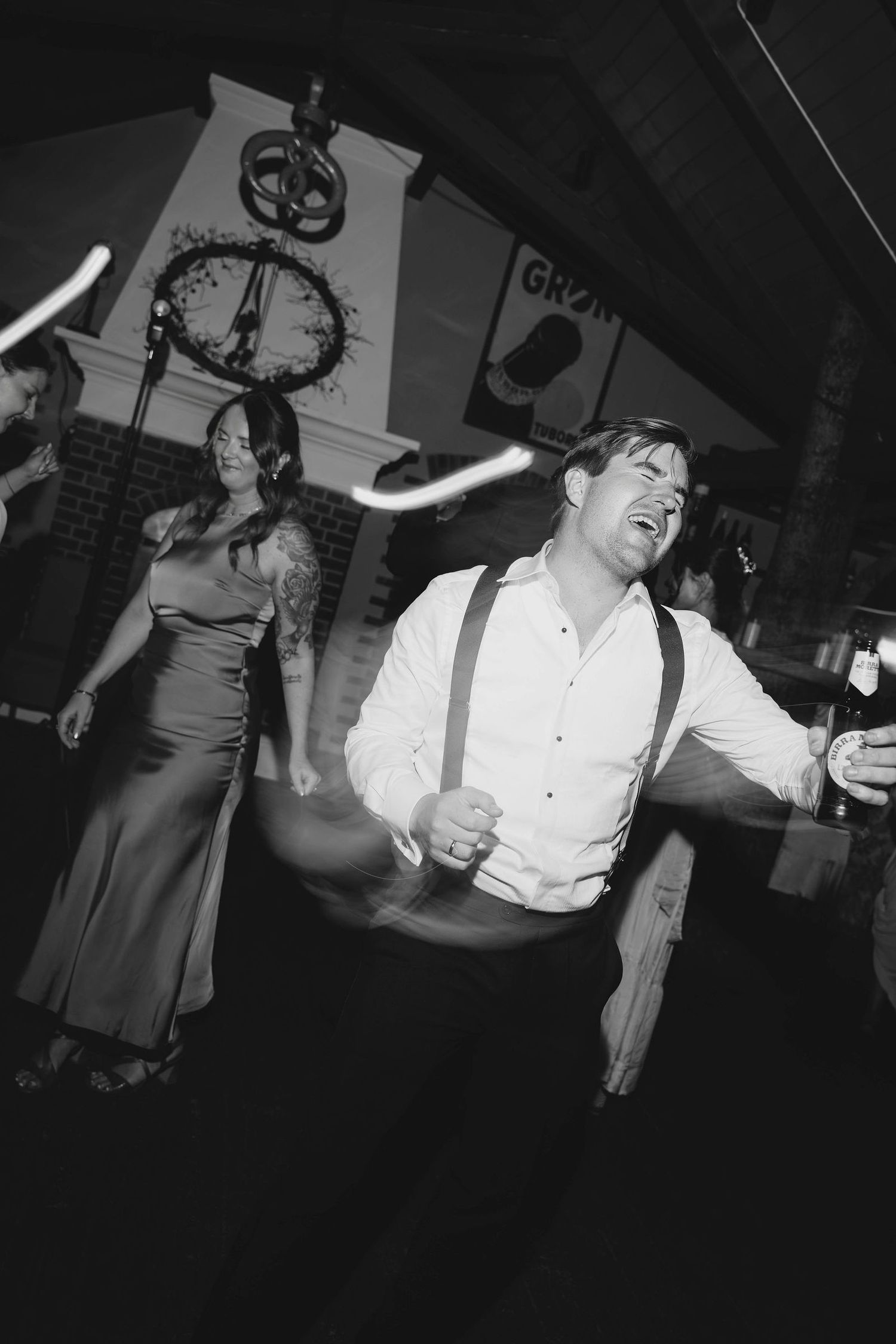 Black and white photo of a man in suspenders dancing energetically at a wedding reception.