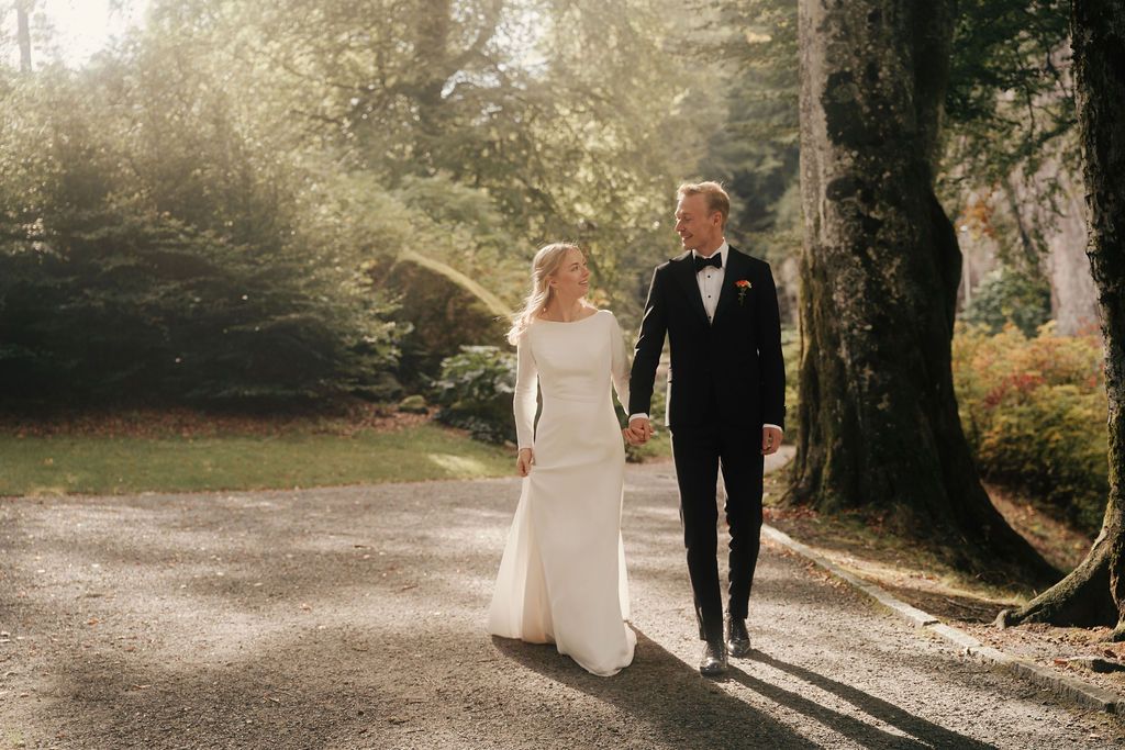 A couple walks hand in hand along a sunlit forest path in formal wedding attire.