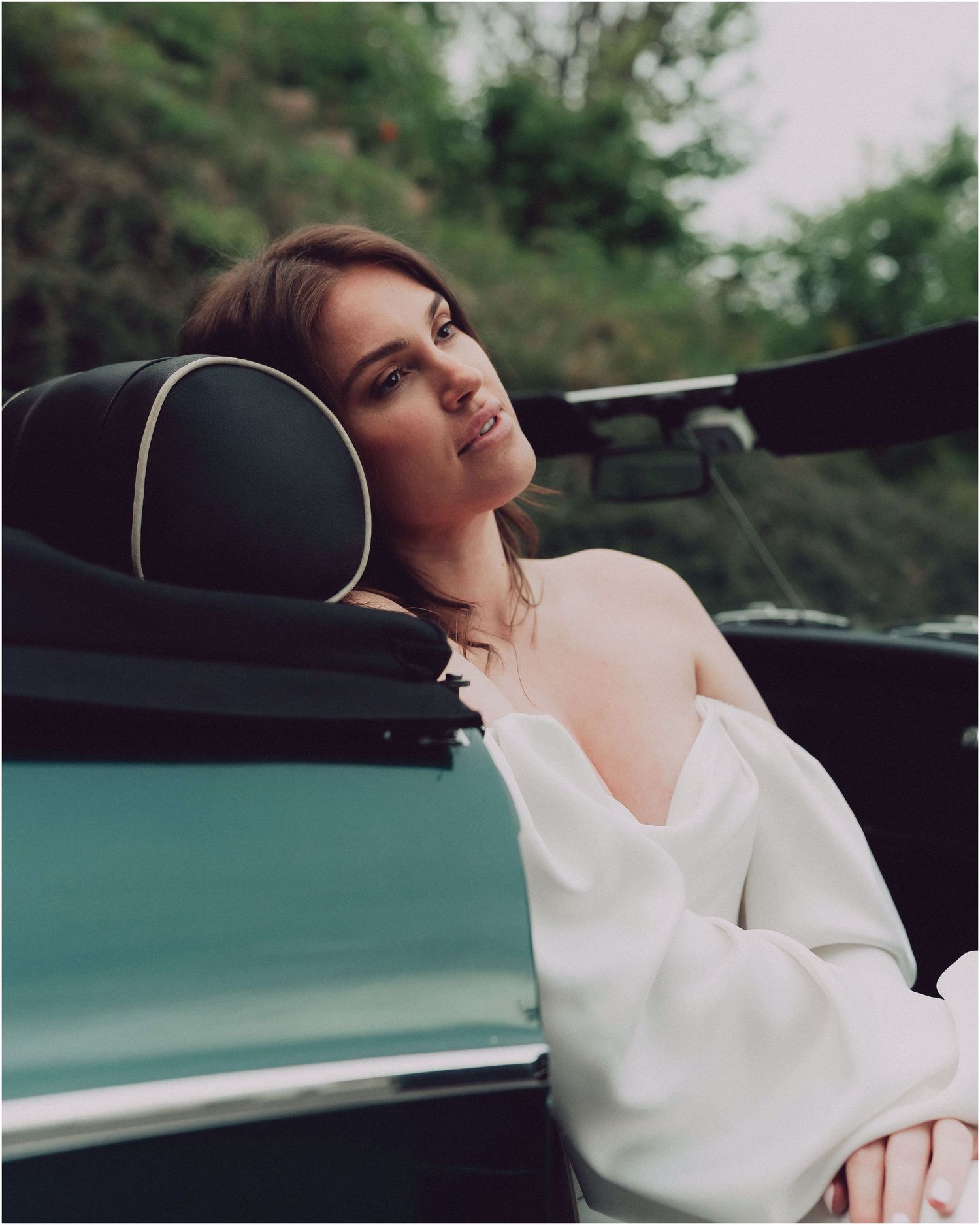 Woman in white sweater reclining in luxury car with teal exterior, captured in moody editorial style photography.