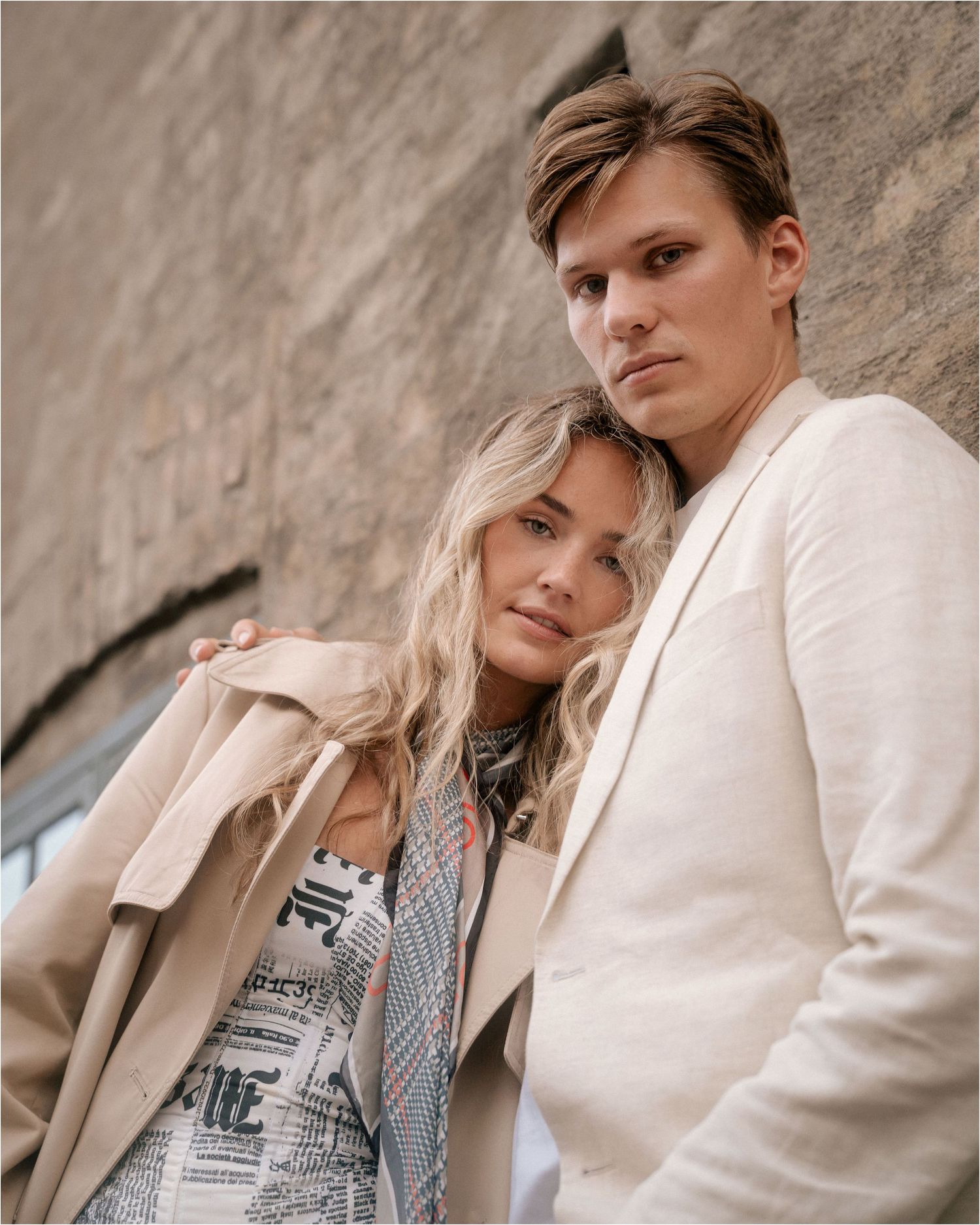 A couple in light-colored clothing poses together against a textured stone wall in an artistic fashion photoshoot.