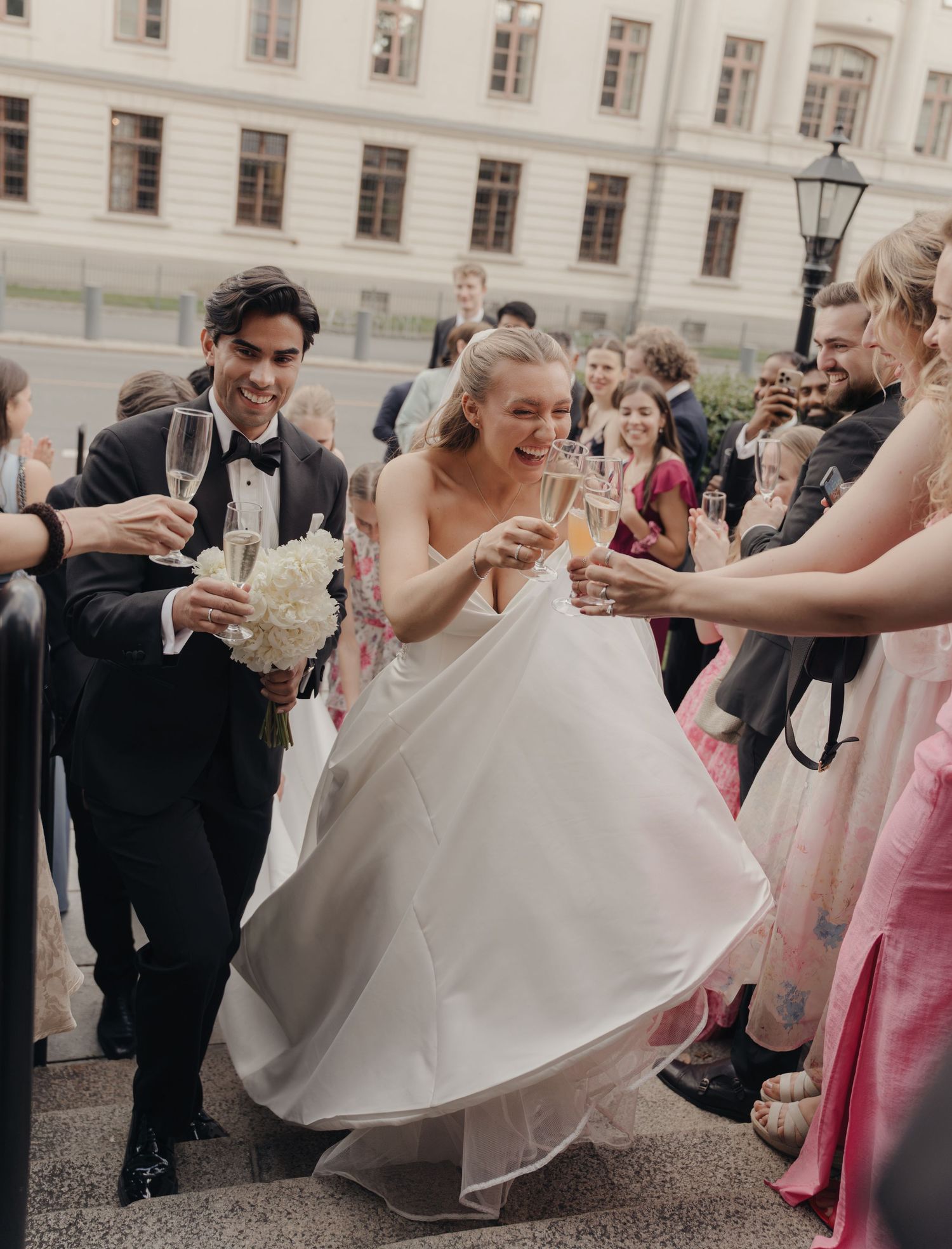 Smiling bride in a white gown and groom holding a bouquet are toasted by cheering wedding guests with champagne glasses.