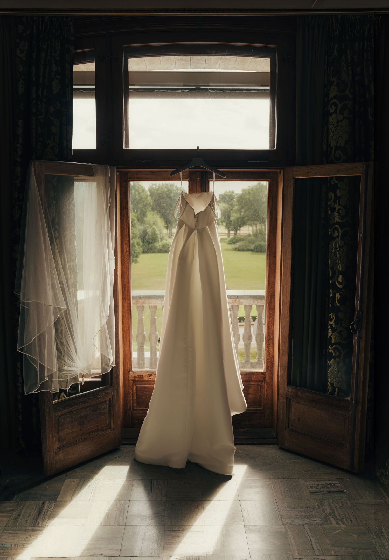 White wedding dress hanging in an open wooden balcony doorway with a sheer veil, sunlight on the floor and a green garden view beyond.
