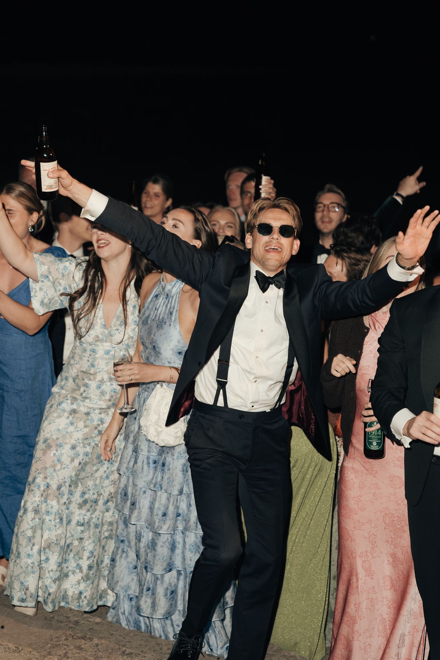 Wedding guests in formal attire dance and cheer with raised arms at night, led by a man in a tuxedo, bow tie and sunglasses.