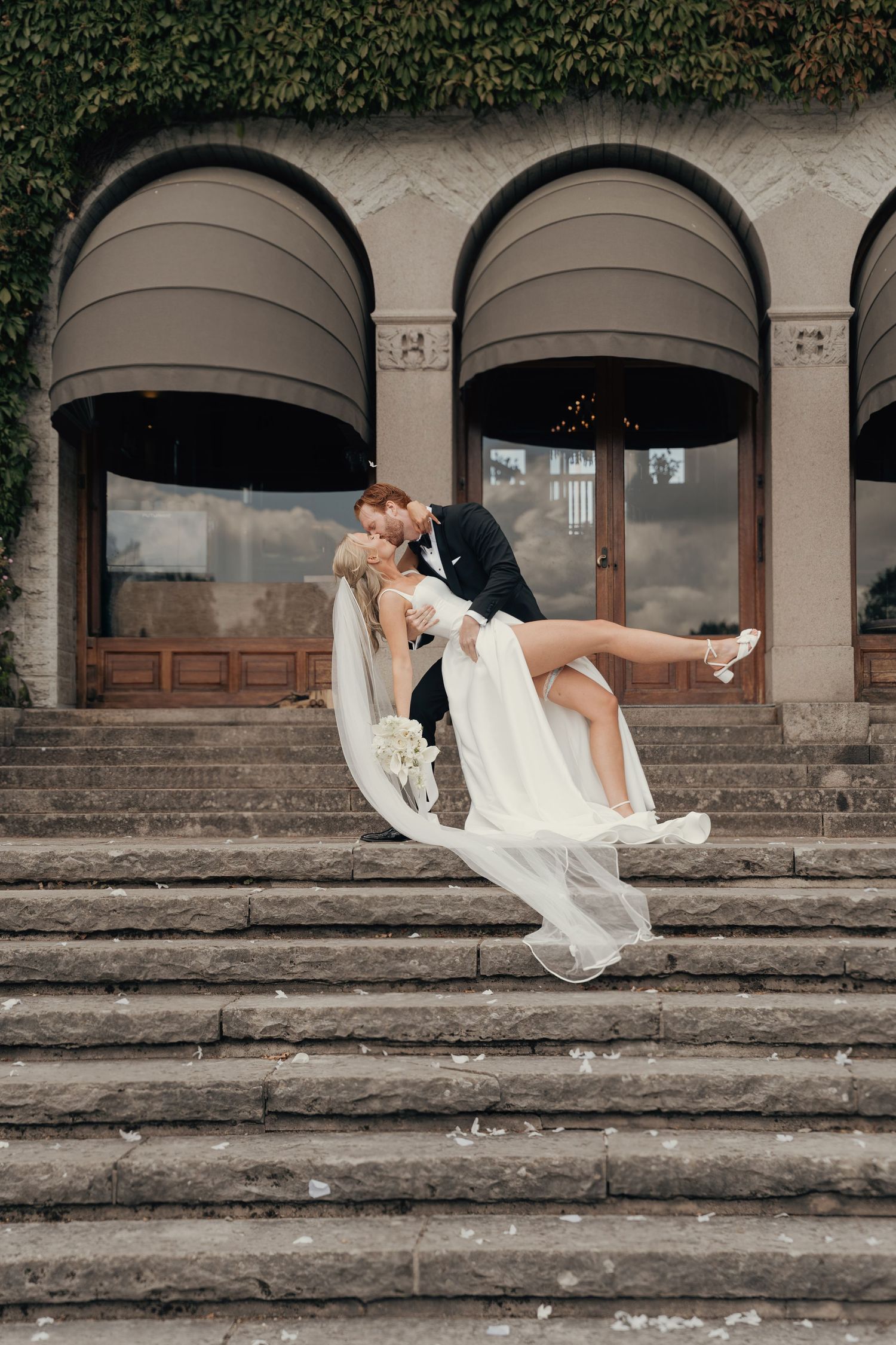 Groom dips bride in a white gown with a flowing veil on grand stone steps in front of an ivy-covered building.
