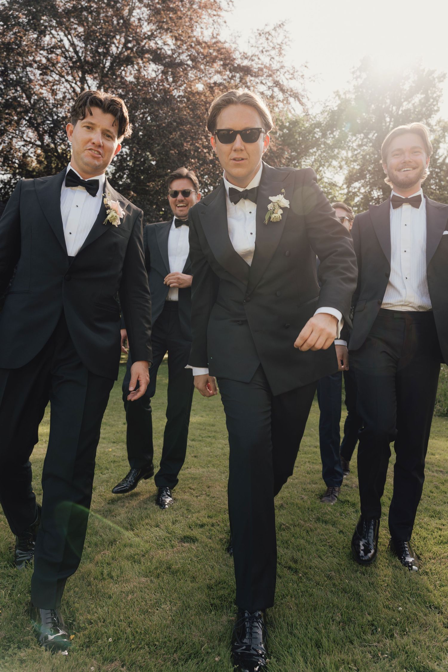 Groomsmen in black tuxedos and bow ties with white boutonnieres walking together across a sunlit lawn at an outdoor wedding.