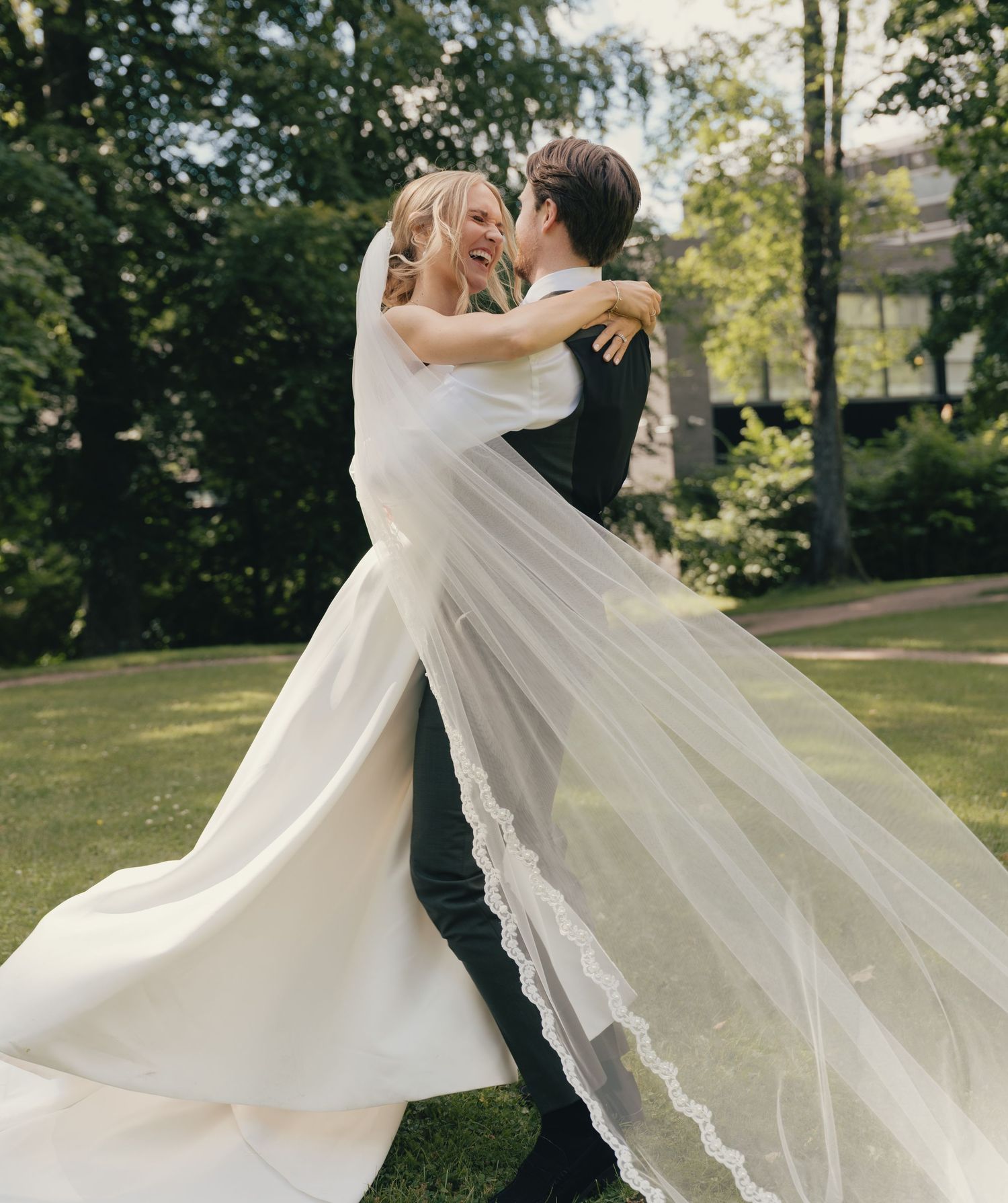 Bride in white gown with lace veil twirling with groom in black suit in sunny garden.