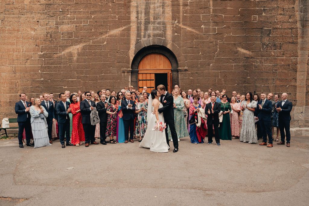 Large wedding party gathered outside historic brick church with arched wooden door for formal group photo.