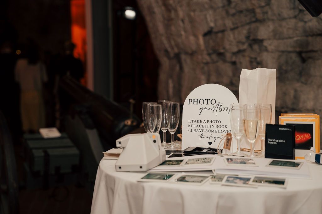 A rustic wedding reception setup with white tablecloths and photo station displays in a dimly lit venue.
