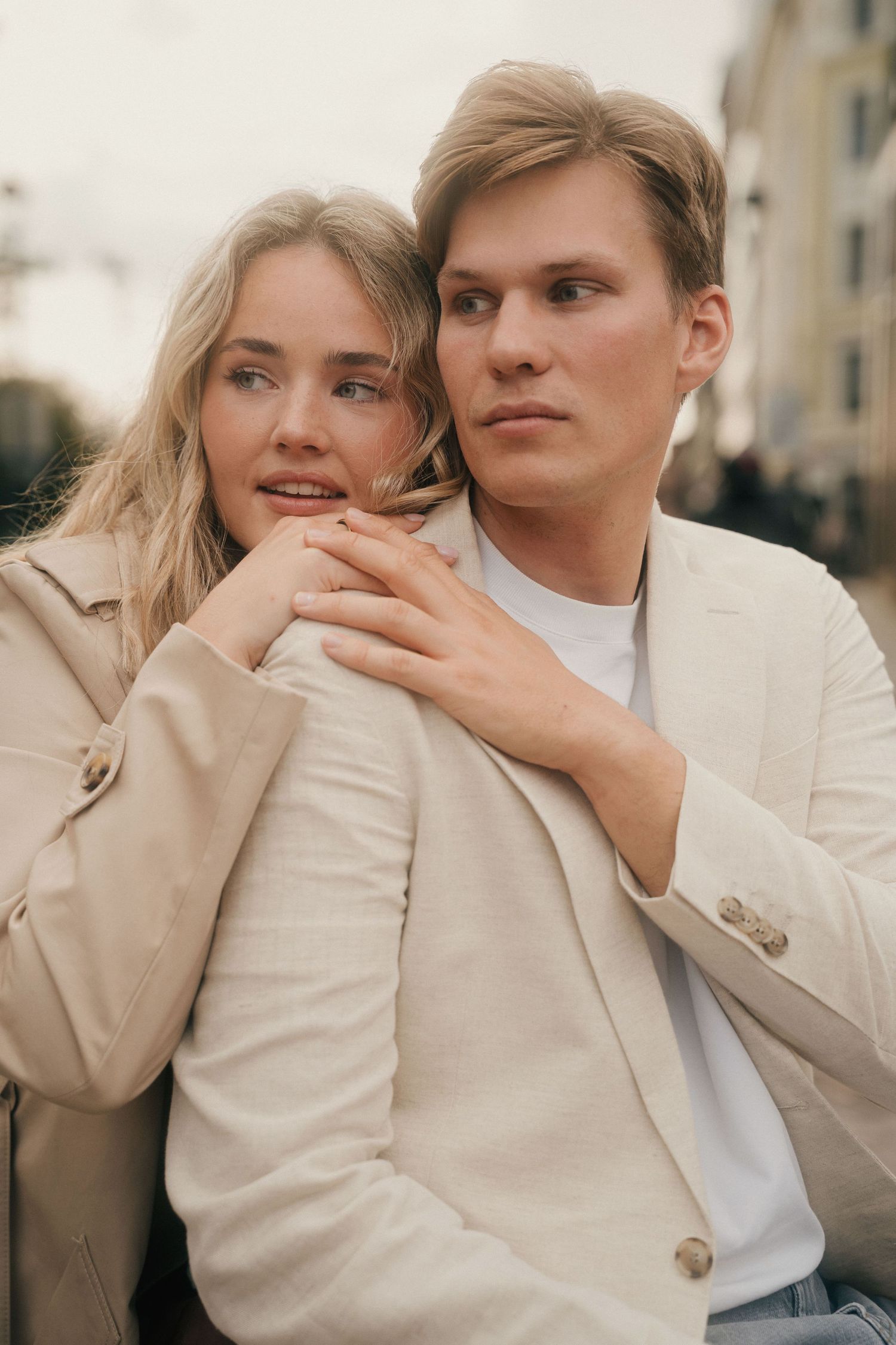 A romantic couple in beige coats embrace outdoors during golden hour with soft lighting and urban backdrop.