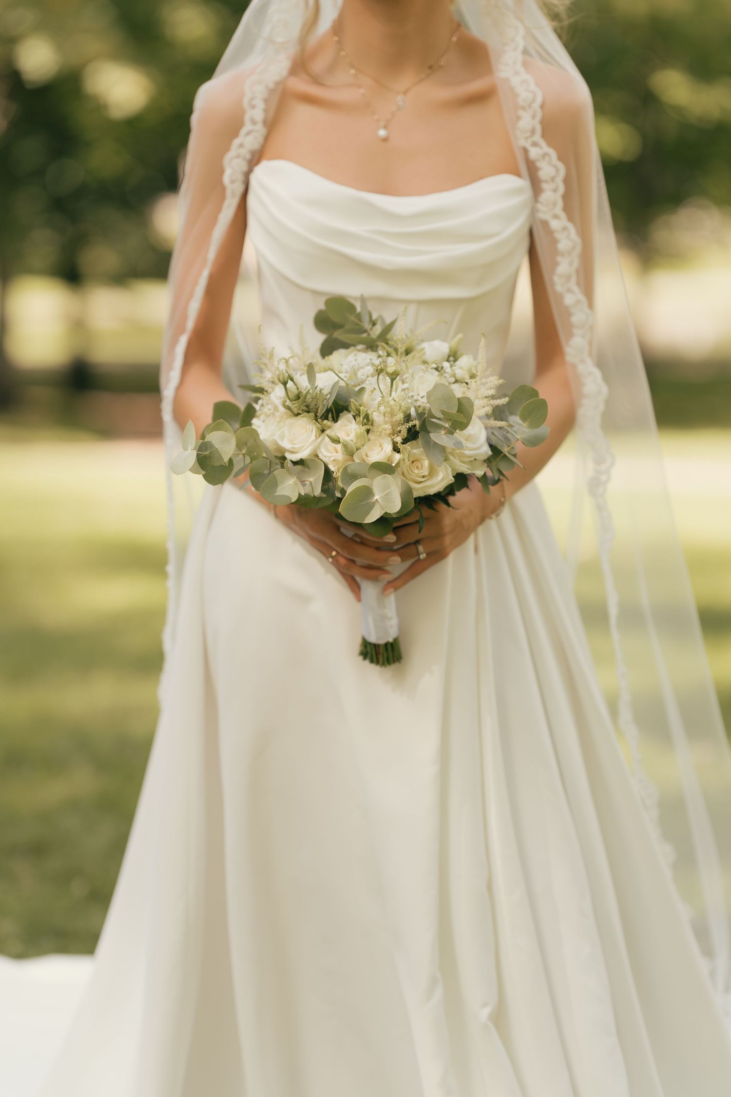 Bride in strapless white gown holding elegant white rose and eucalyptus bouquet with lace veil outdoors.