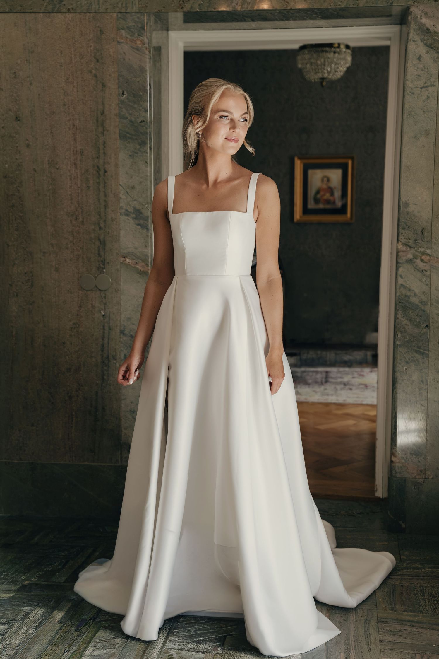 Bride in an elegant white satin A-line wedding gown with square neckline and straps stands in a marble hallway with a framed portrait.