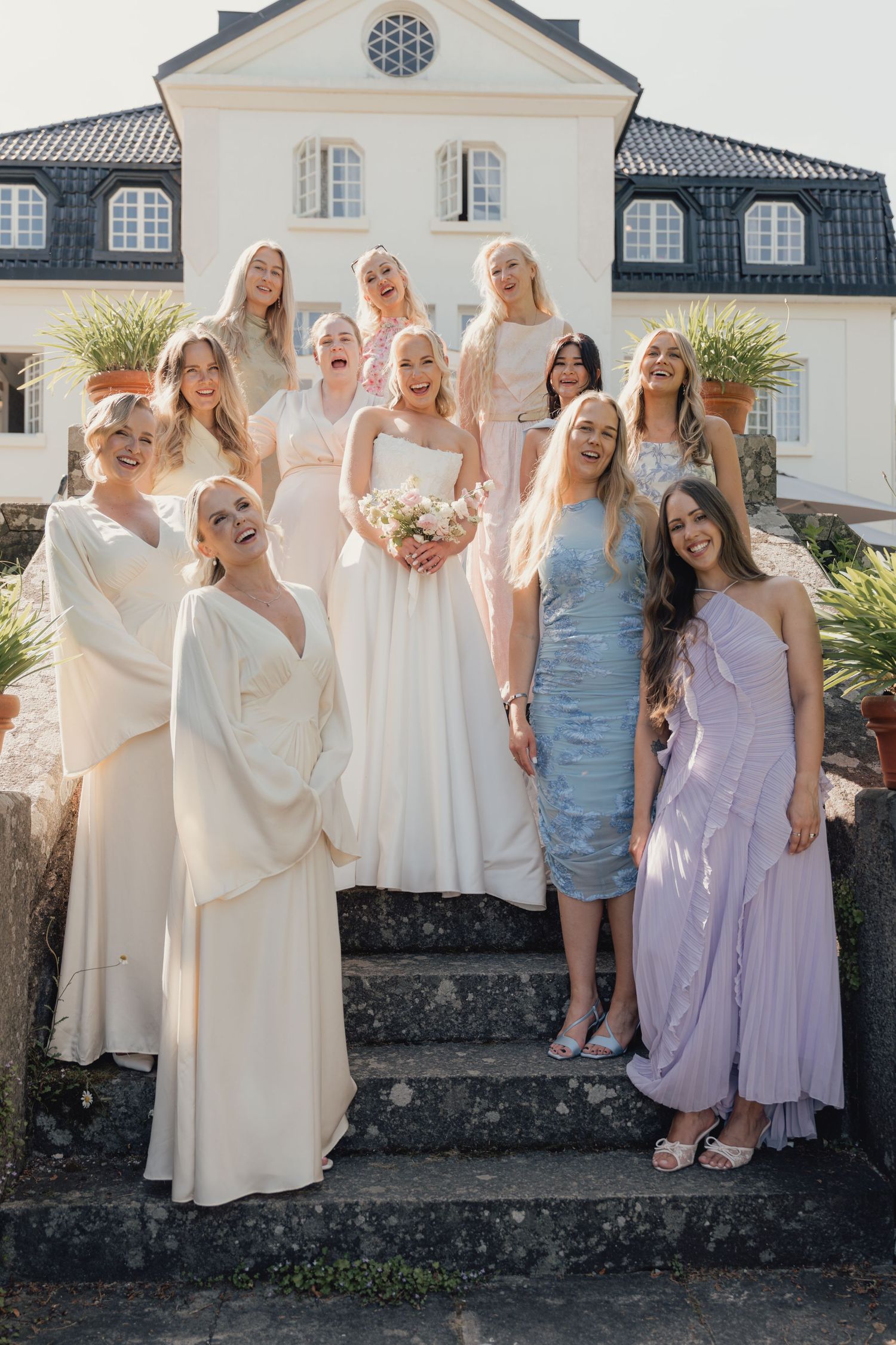 Bride in a white gown stands on stone steps with smiling bridesmaids in cream and pastel dresses before a white manor.