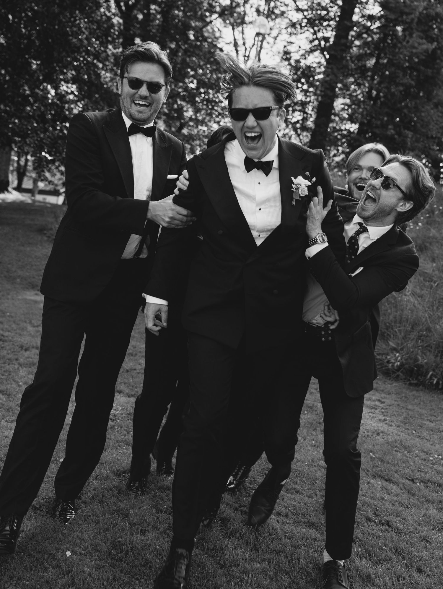 Black and white photo of groomsmen in tuxedos and sunglasses laughing as they playfully lift the groom outdoors on a grassy lawn.