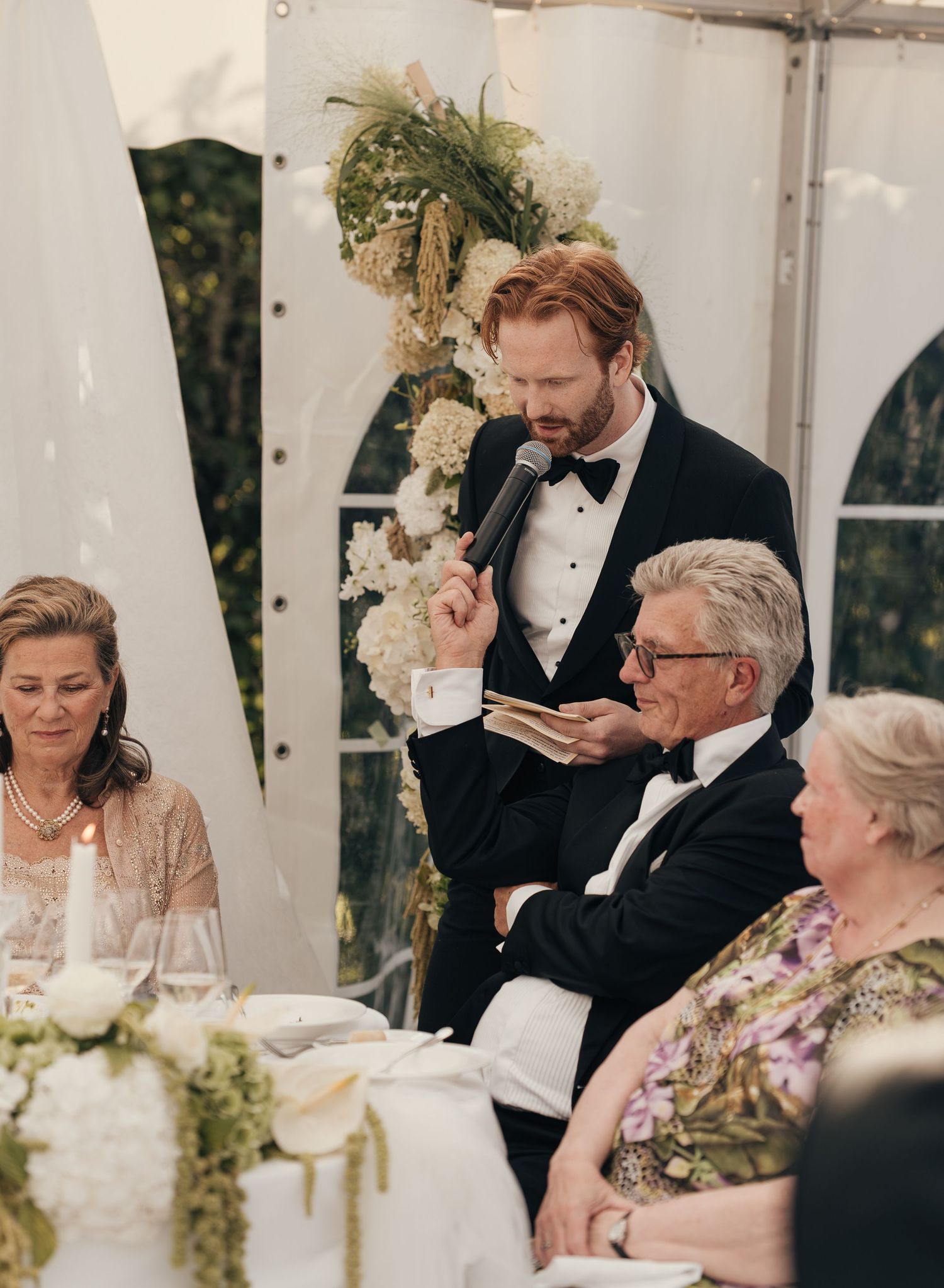 A bearded man in a tuxedo gives a wedding toast with a microphone as seated guests listen under a white tent.
