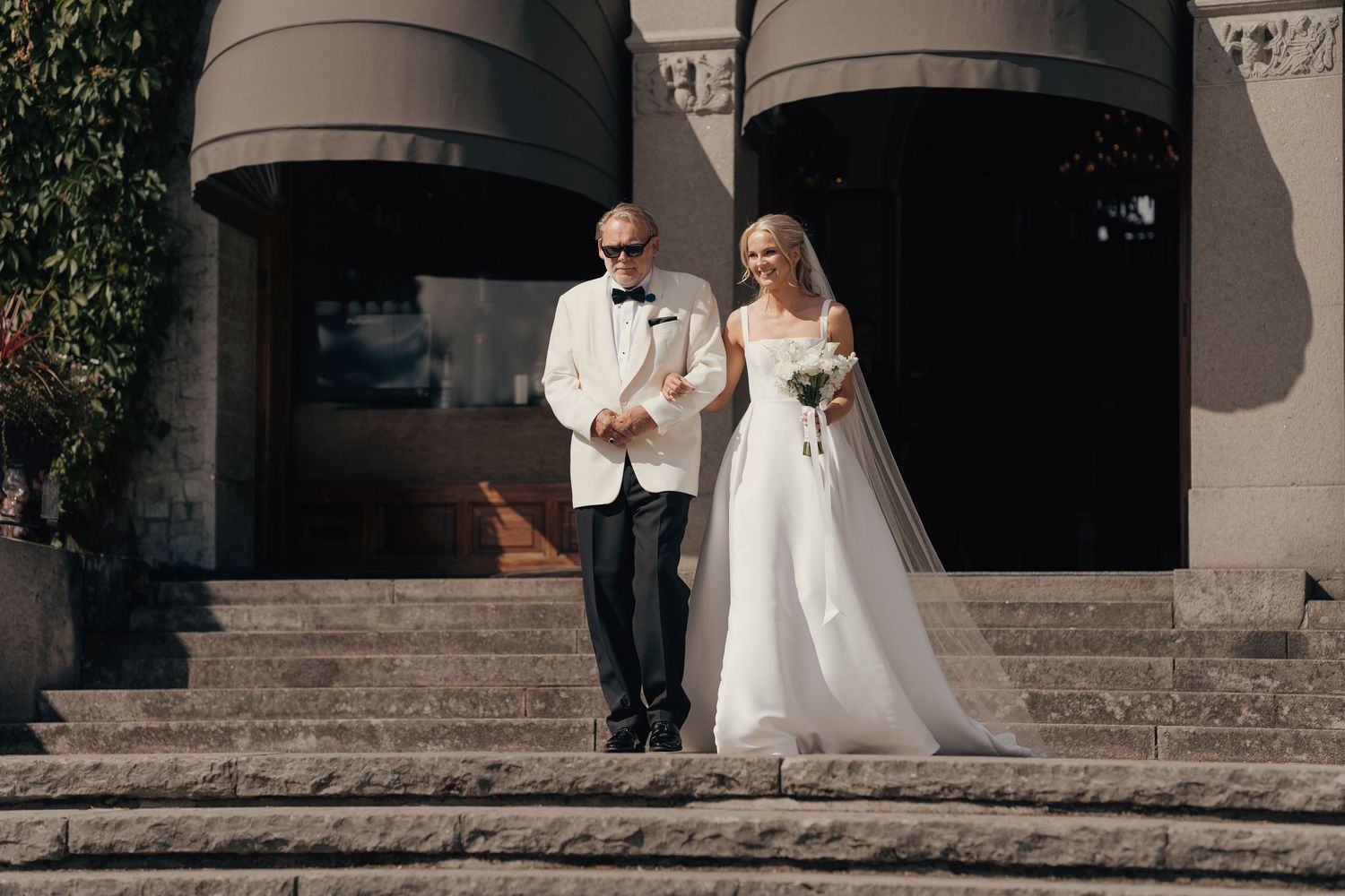 A bride in a white gown and veil holding a bouquet walks down stone steps arm in arm with a man in a white dinner jacket and bow tie.