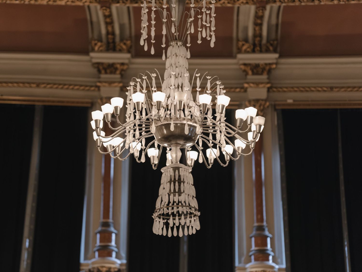 Ornate crystal chandelier with white glass shades hanging in an elegant hall with dark curtains and decorative columns.