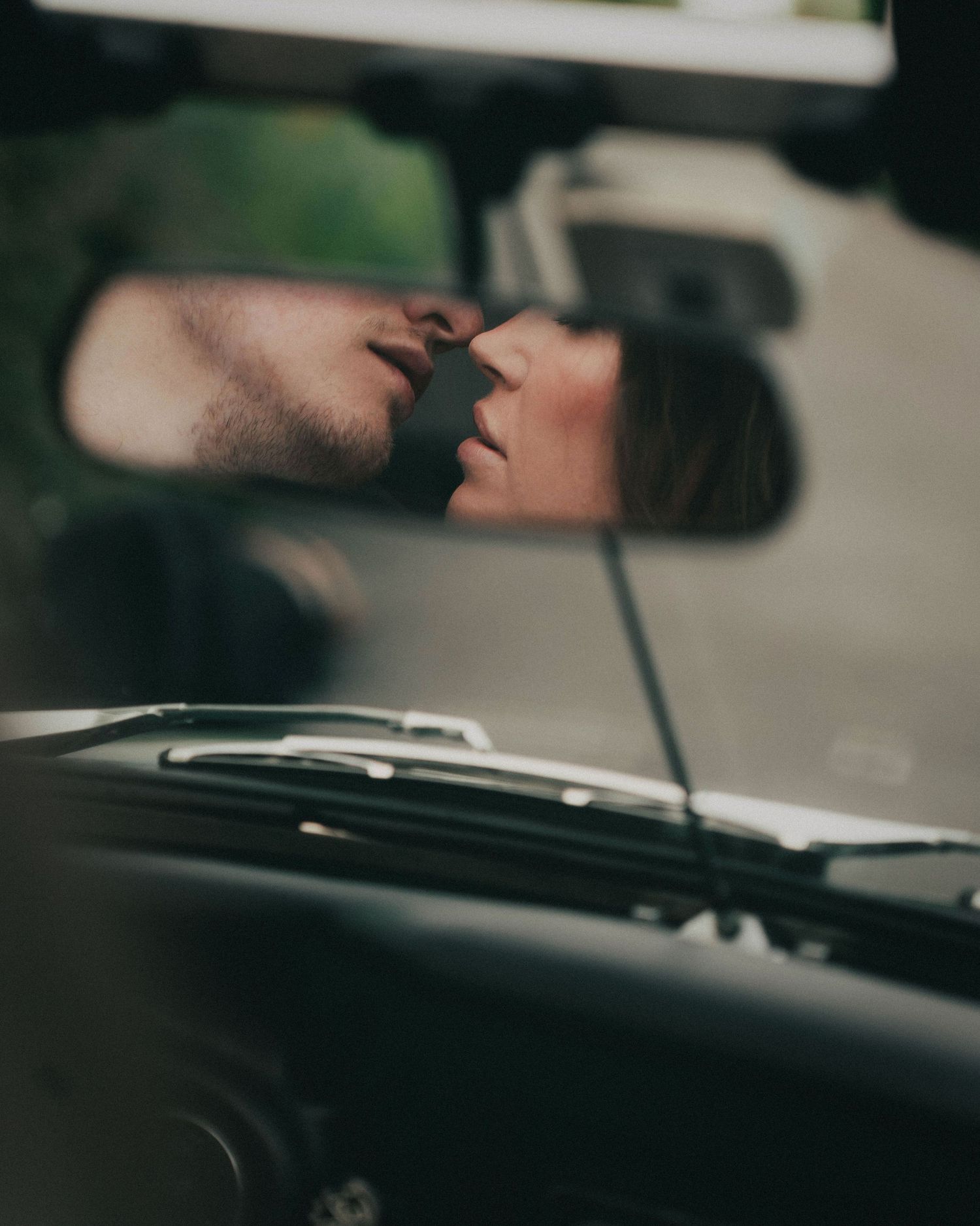 A romantic moment reflected in a car's rearview mirror, with the dashboard and windshield visible in the foreground.