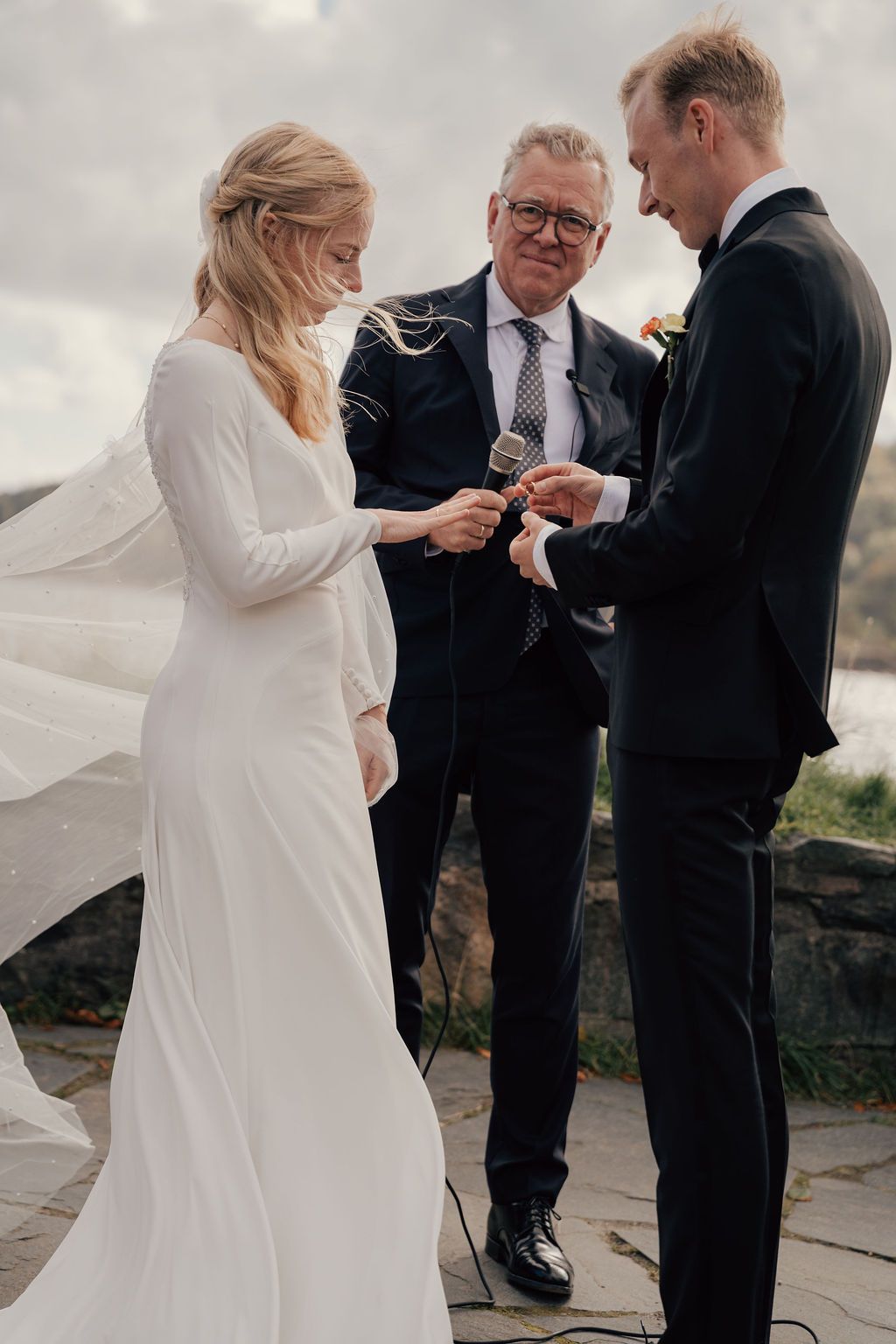 A couple exchanges rings during an outdoor wedding ceremony with an officiant present.