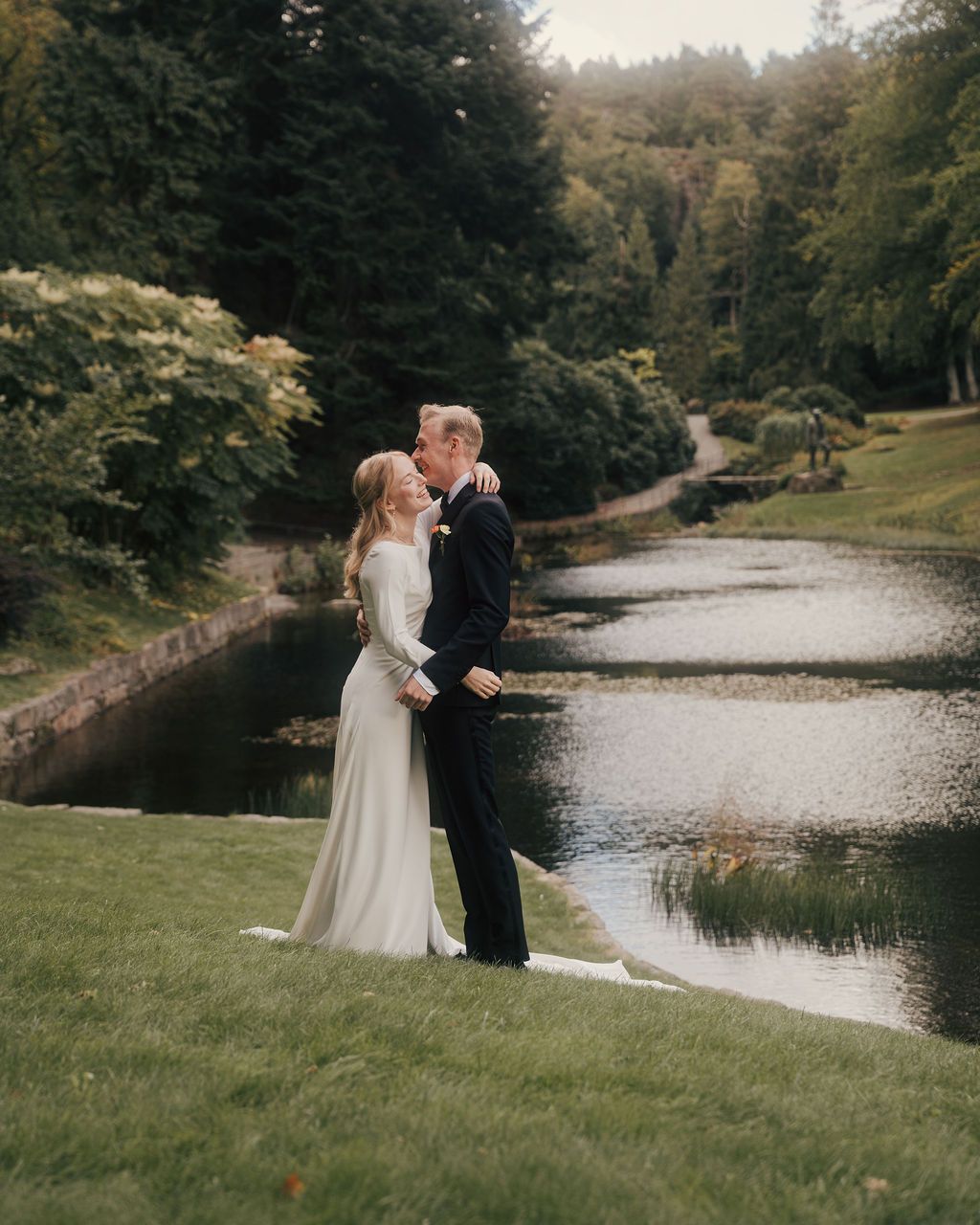 A couple shares an intimate moment by a serene lake surrounded by autumn trees and natural scenery.