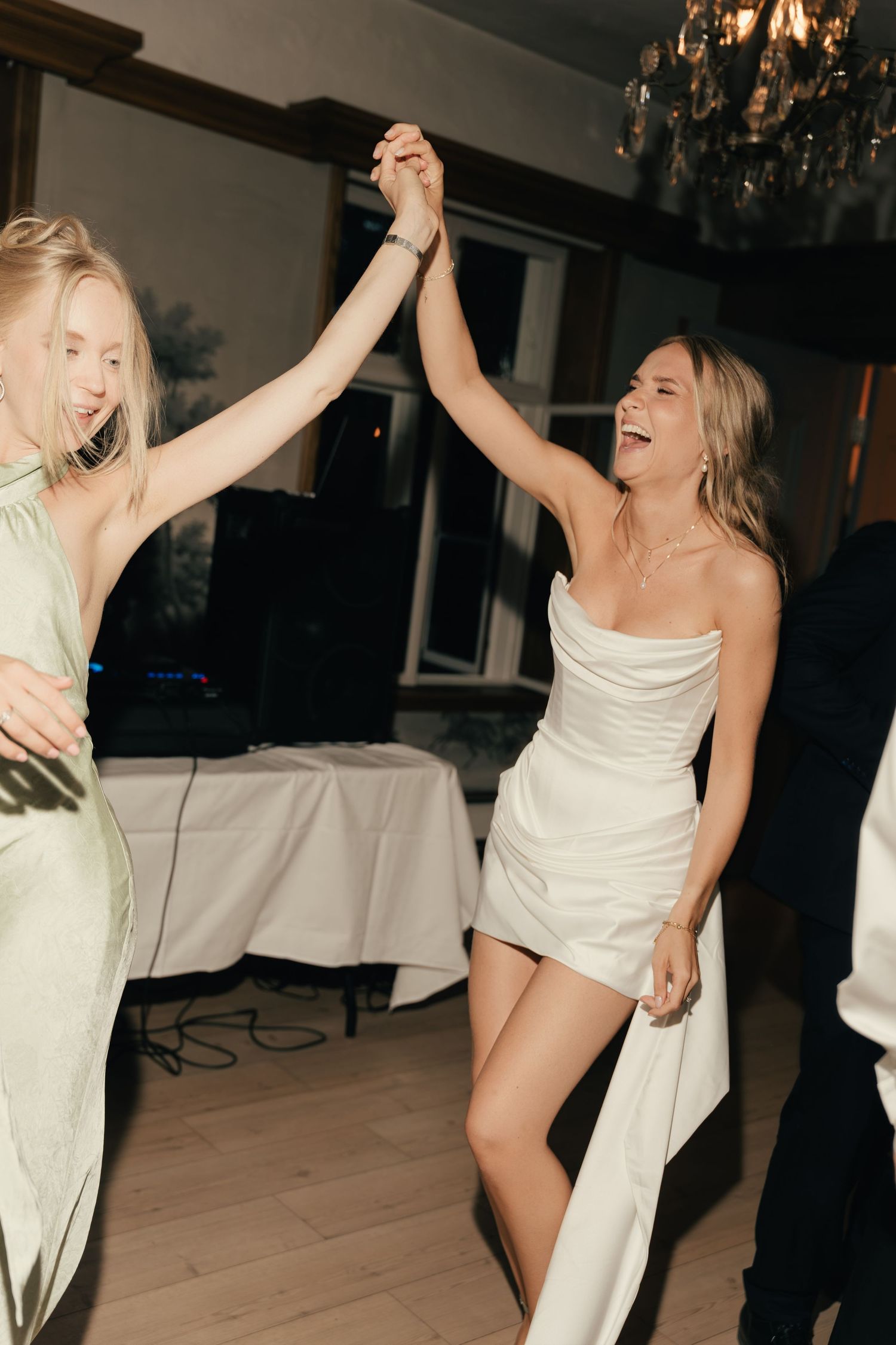 Bride in short white dress dancing and holding hands with bridesmaid at wedding reception.