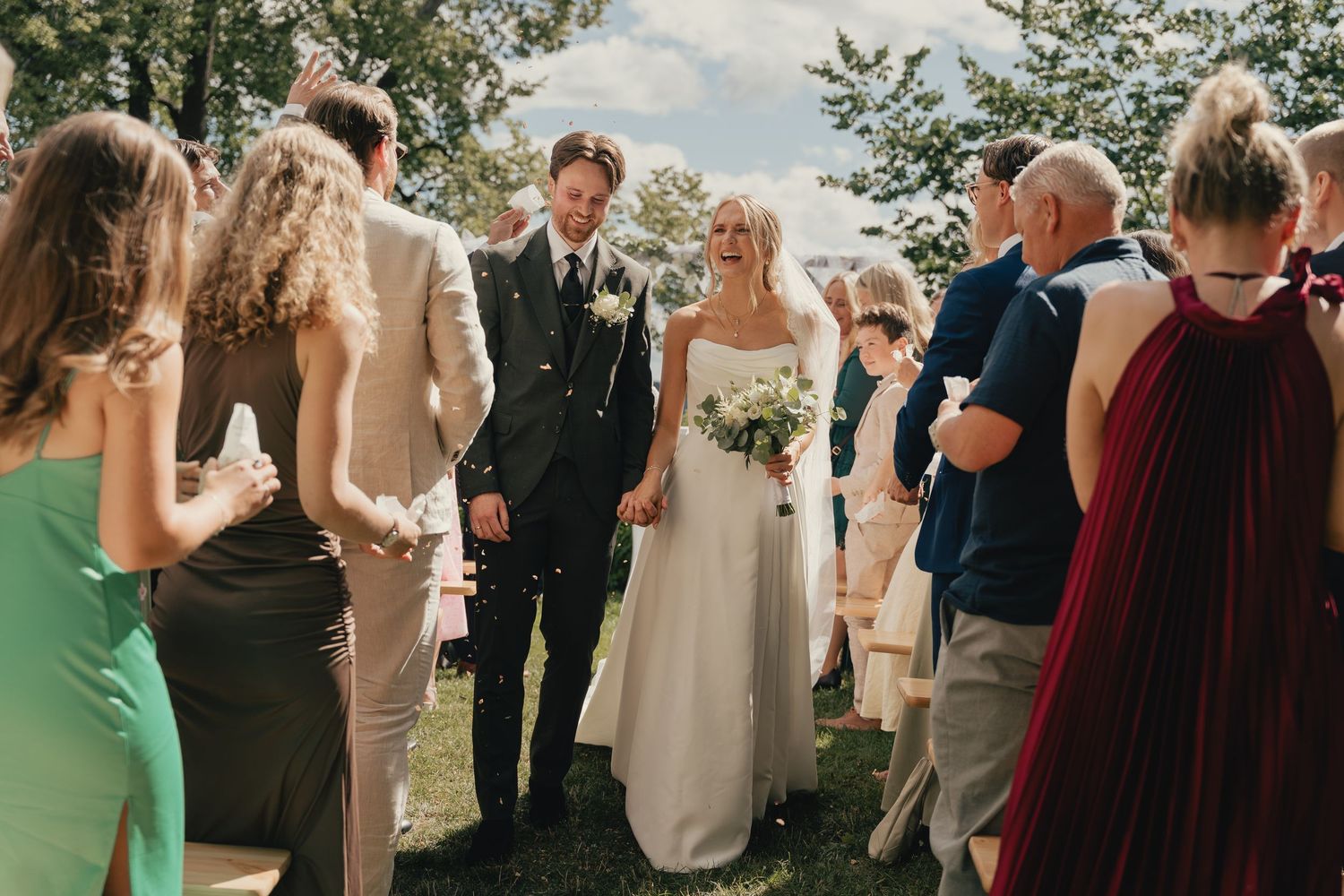 Bride and groom walk down the aisle at an outdoor wedding ceremony surrounded by smiling guests.