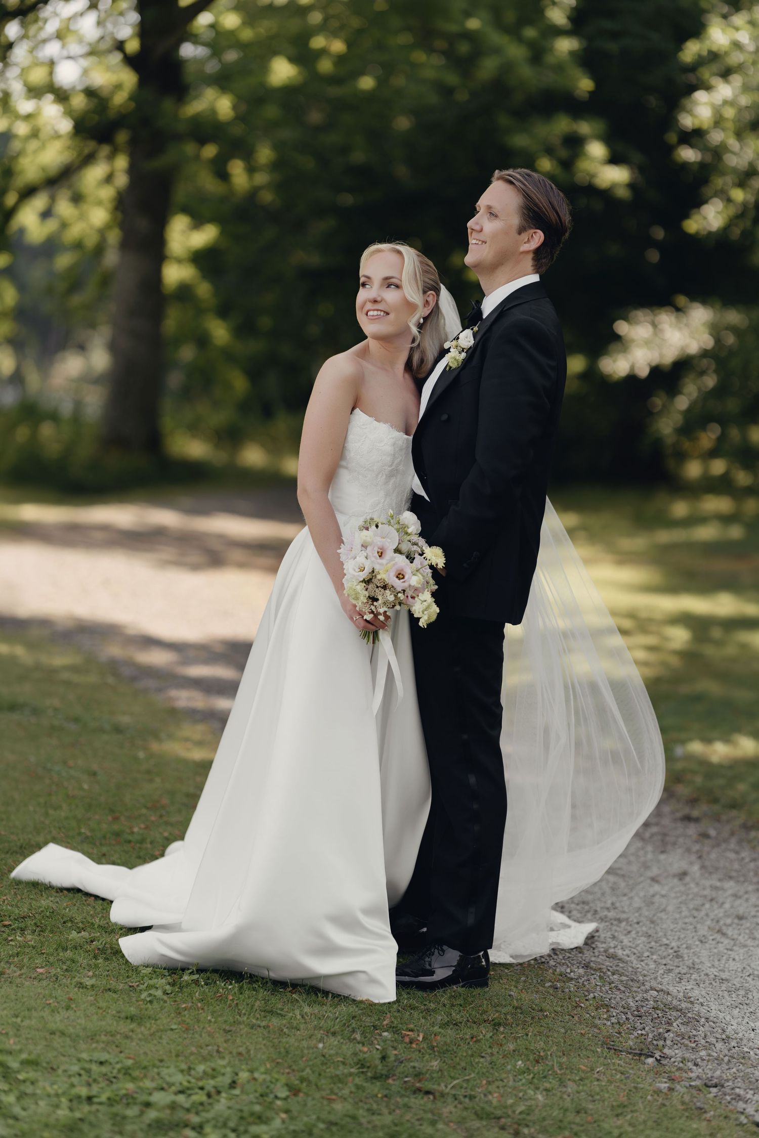 Bride in a strapless white gown holding a bouquet stands beside her groom in a black tuxedo on a tree-lined path.