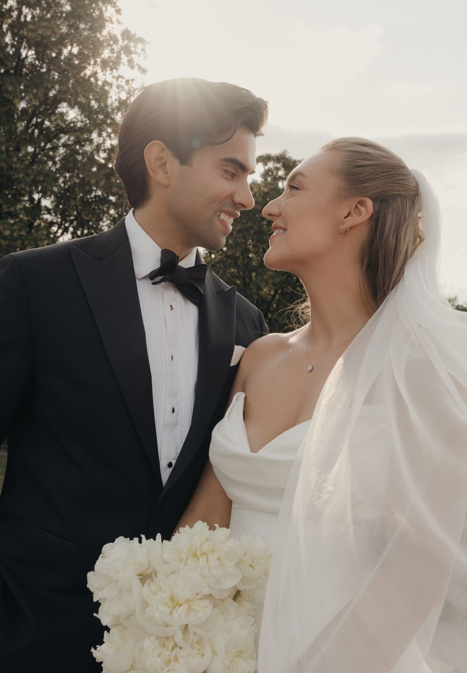 Smiling bride in a white strapless gown and veil holds a white peony bouquet, gazing at her groom in a black tuxedo and bow tie.