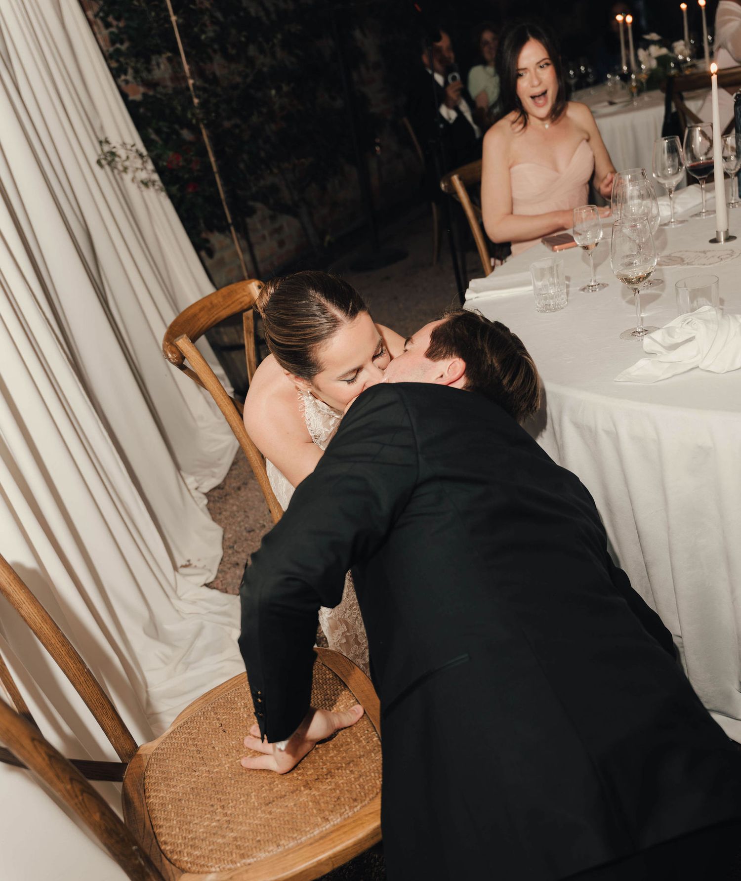 Couple sharing a romantic kiss at a wedding reception while a guest reacts with joy in the background.