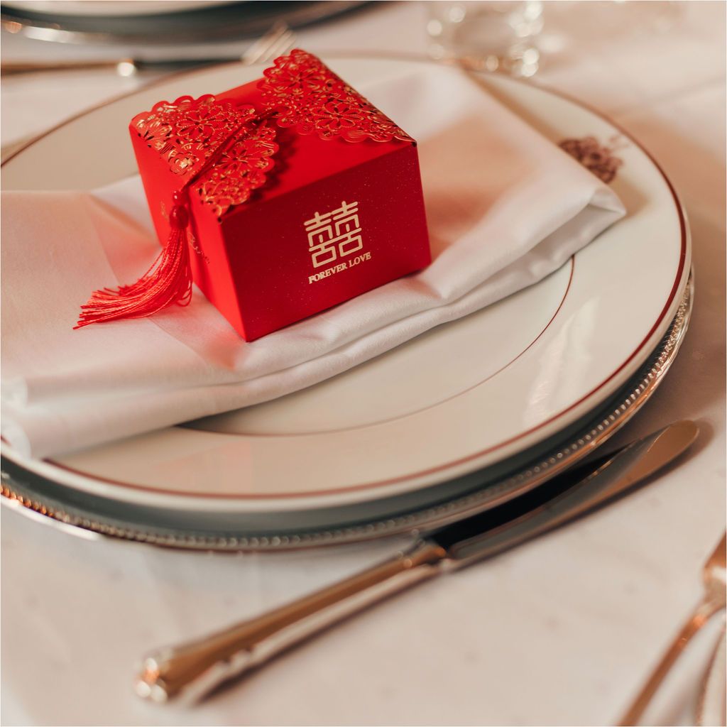 A red Chinese wedding favor box with tassel sits elegantly on a white plate with silver trim and chopsticks.