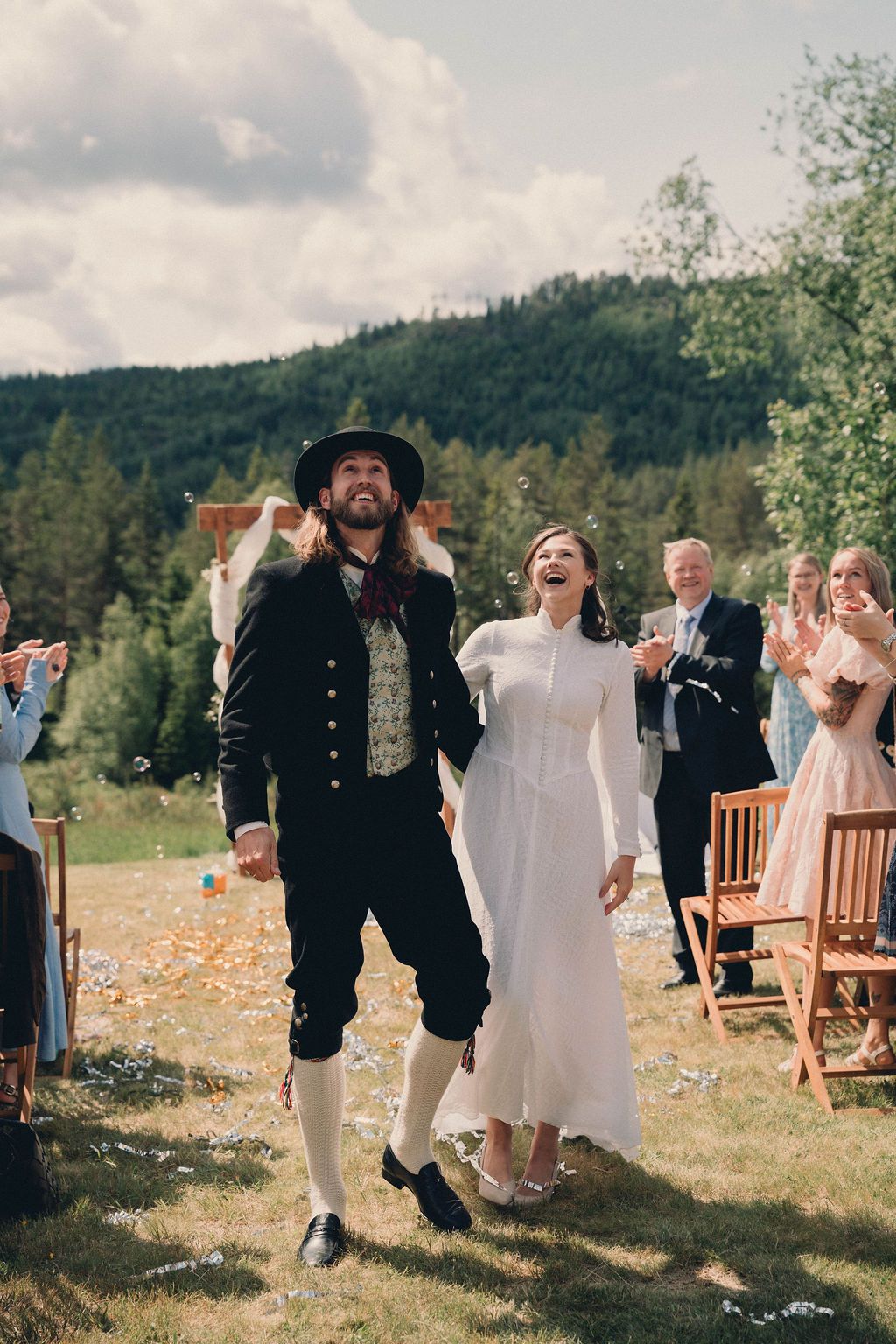 A couple celebrates during an outdoor wedding ceremony in traditional folk attire against a mountainous forest backdrop.