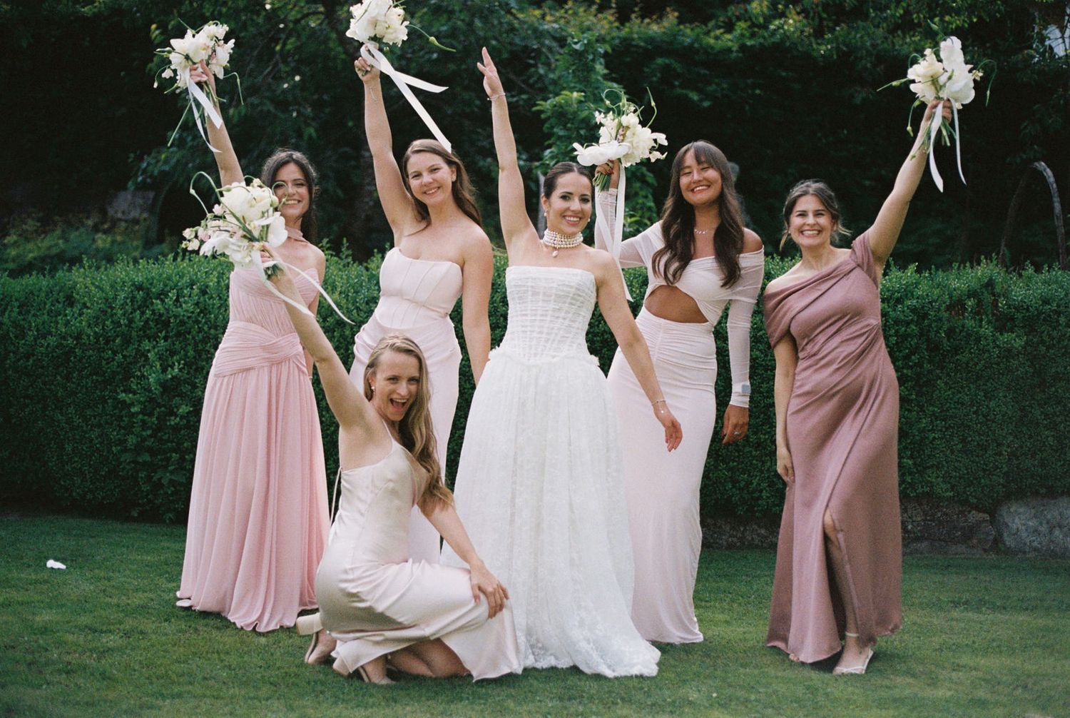 Bride in white gown poses with five bridesmaids in pink and mauve dresses, raising bouquets outdoors on green lawn.