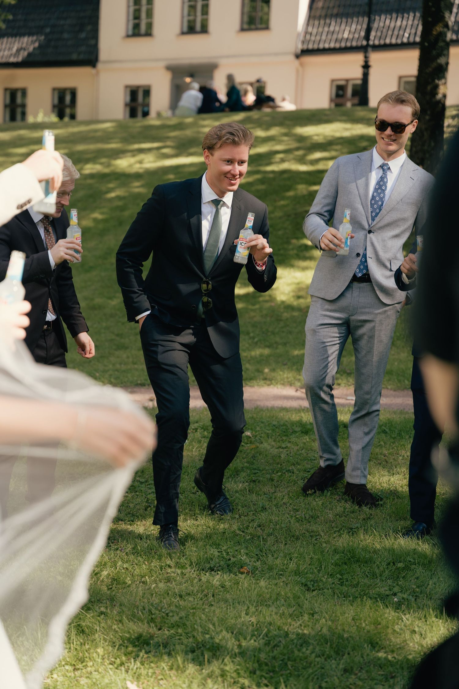 Smiling wedding guests in suits celebrate outdoors with champagne glasses on green lawn.