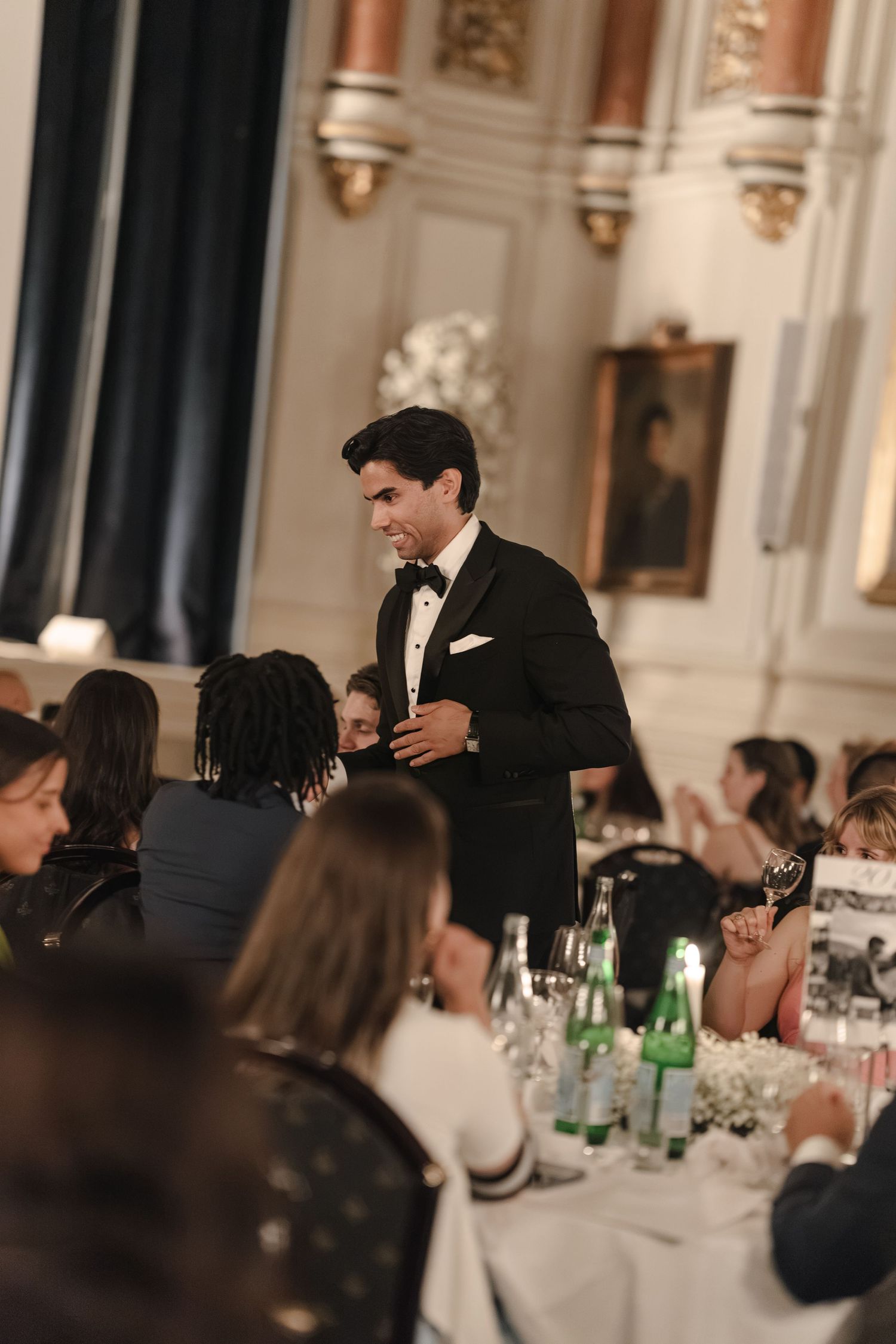Man in a black tuxedo and bow tie stands among seated guests at a formal banquet in an ornate hall with gilded details.