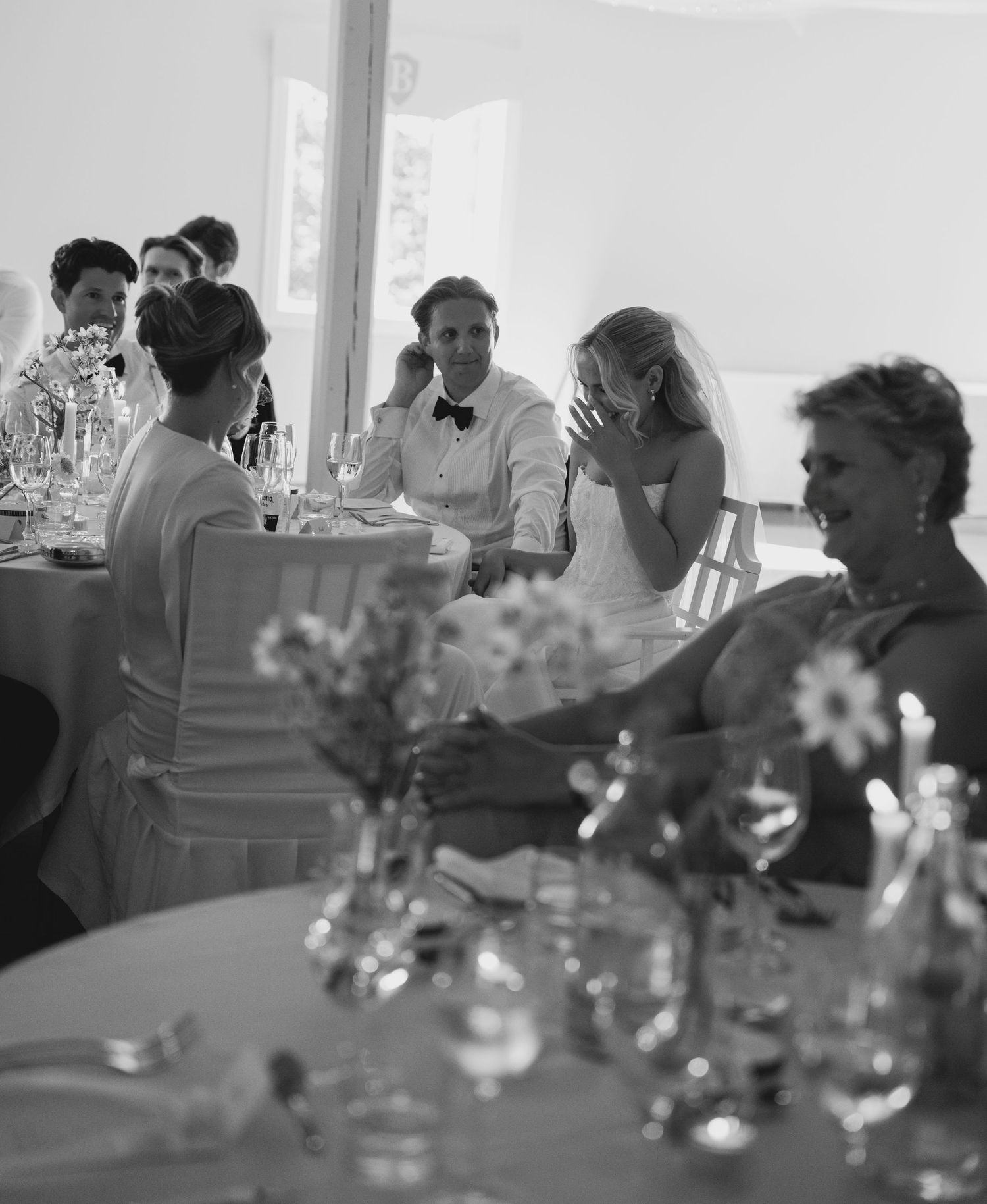 Black and white wedding reception photo of a bride laughing with her hand over her face beside the smiling groom at a table.