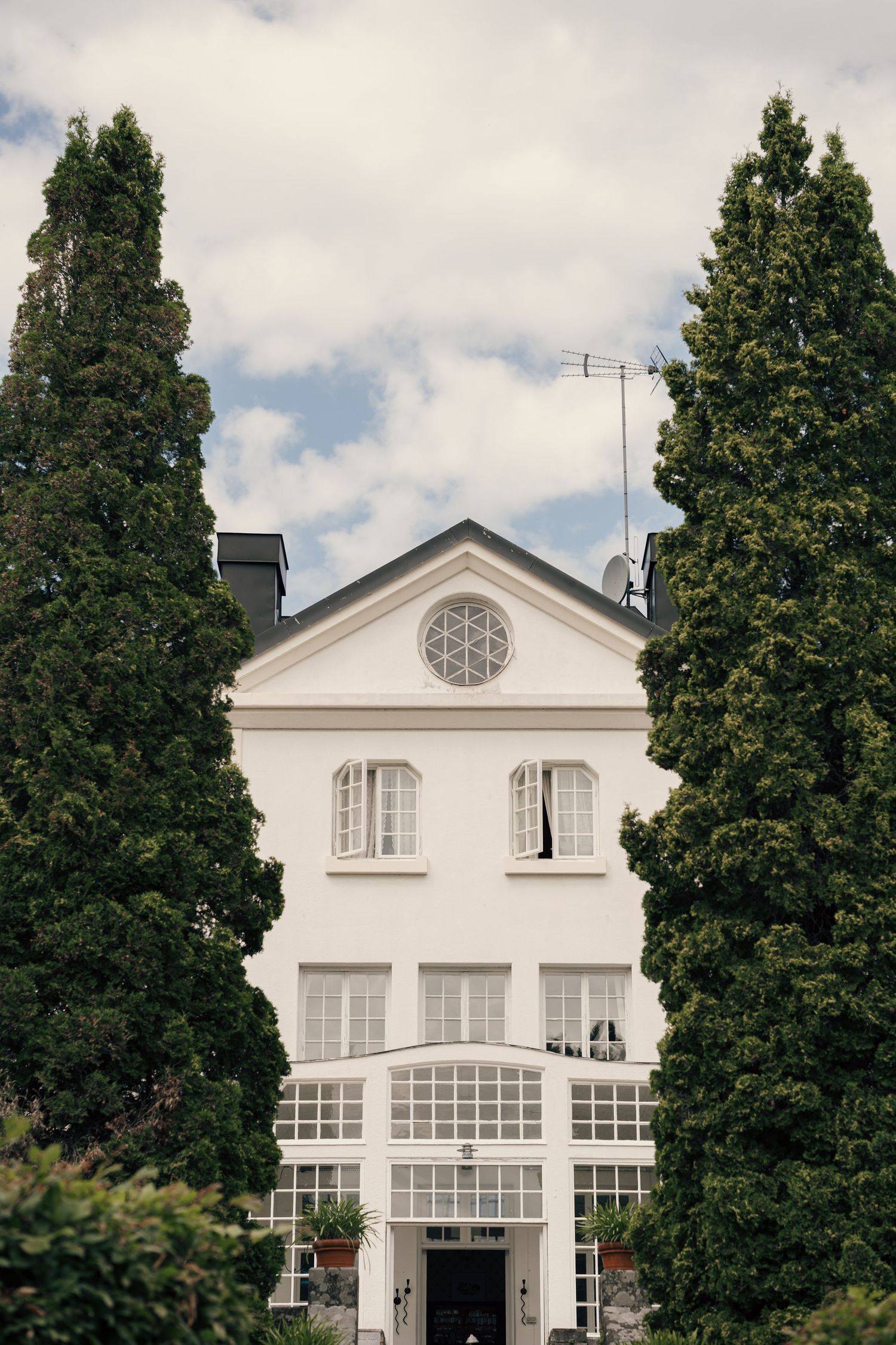 White neoclassical mansion with a circular gable window and tall windows, framed by two tall evergreen trees under a cloudy sky.