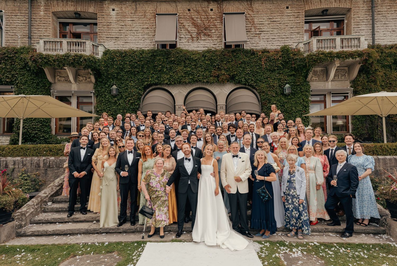 Large wedding party in formal attire poses with the bride in a white gown on stone steps in front of an ivy-covered building.