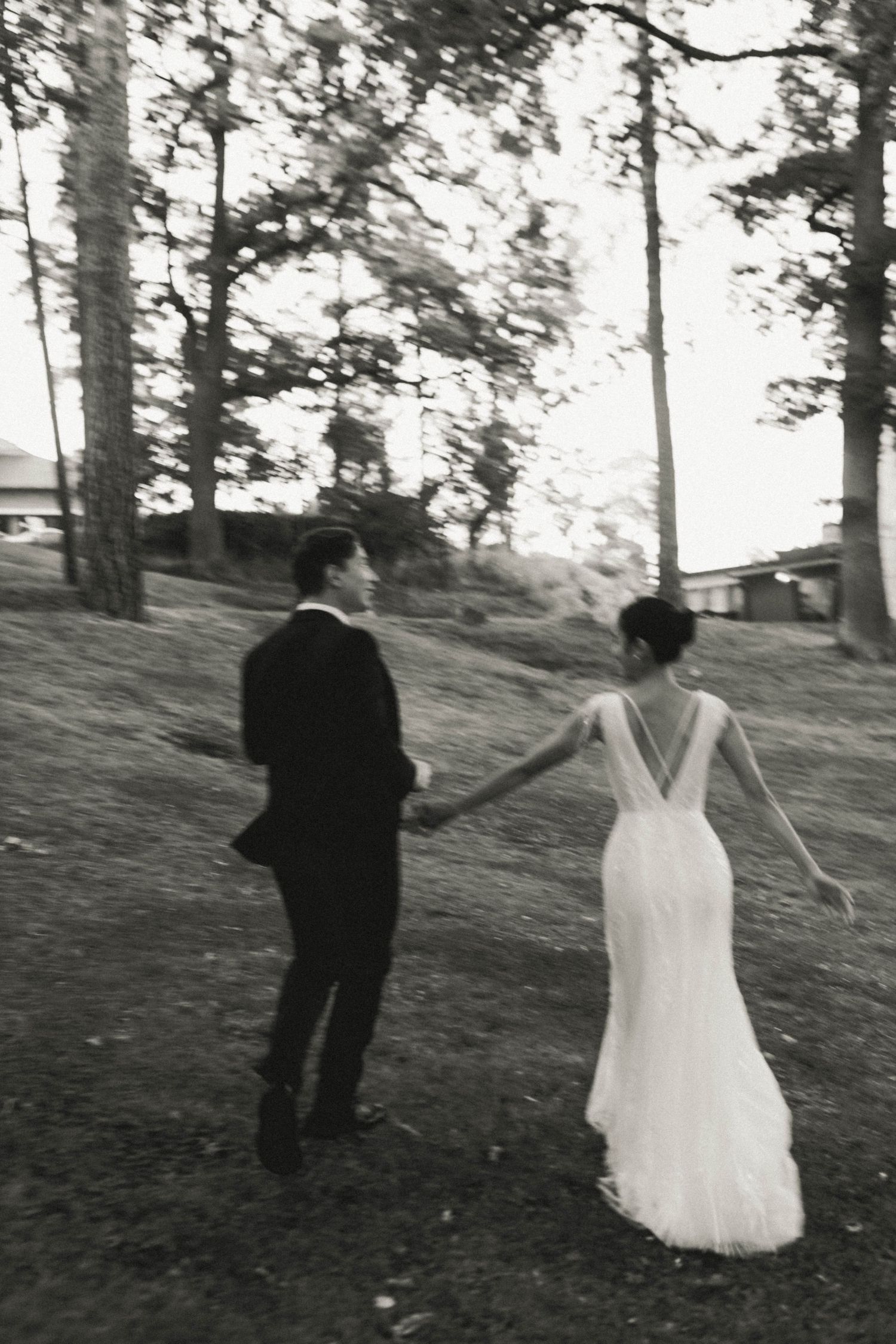 A couple in wedding attire walks hand in hand on a hillside among tall trees in black and white photography.