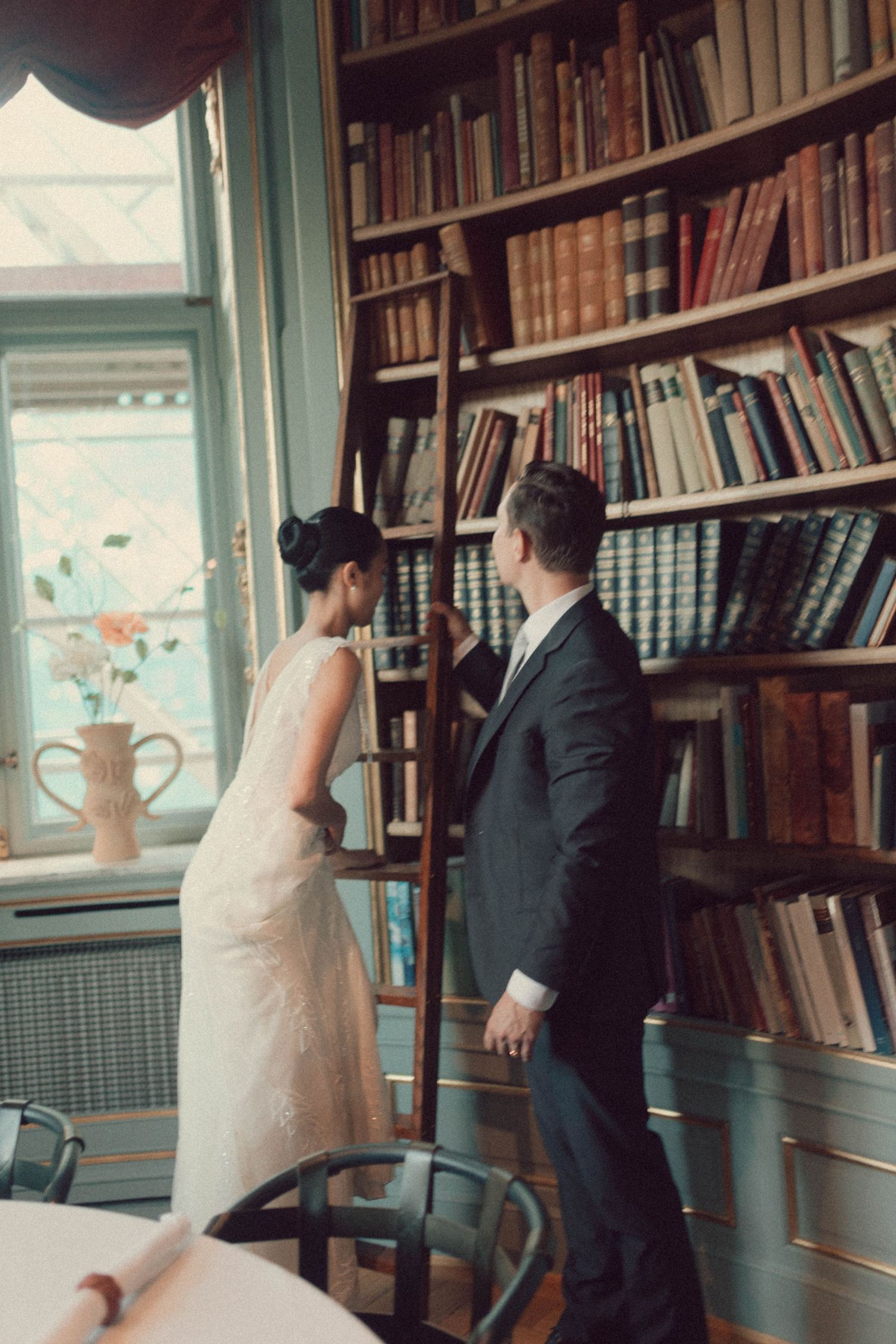 A couple in wedding attire share an intimate moment in a vintage library setting with floor-to-ceiling bookshelves.